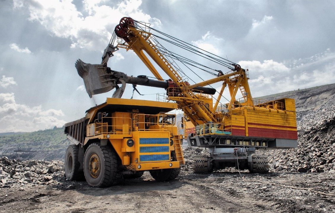 L'image présente un site d'extraction minière. Au premier plan, un gros camion de transport de couleur jaune, avec des roues massives, est chargé de matériaux. À côté, une grande pelle mécanique jaune et rouge, dotée d'un bras articulé, a relevé une charge de terre ou de roches. Le paysage environnant est accidenté, avec des tas de gravier et des pentes abruptes. Le ciel est partiellement nuageux, laissant passer des rayons de lumière qui illuminent la scène, créant une ambiance industrielle dynamique. Des bruits de machines et d'activités minières peuvent être imaginés, rendant l'environnement à la fois impressionnant et robuste.