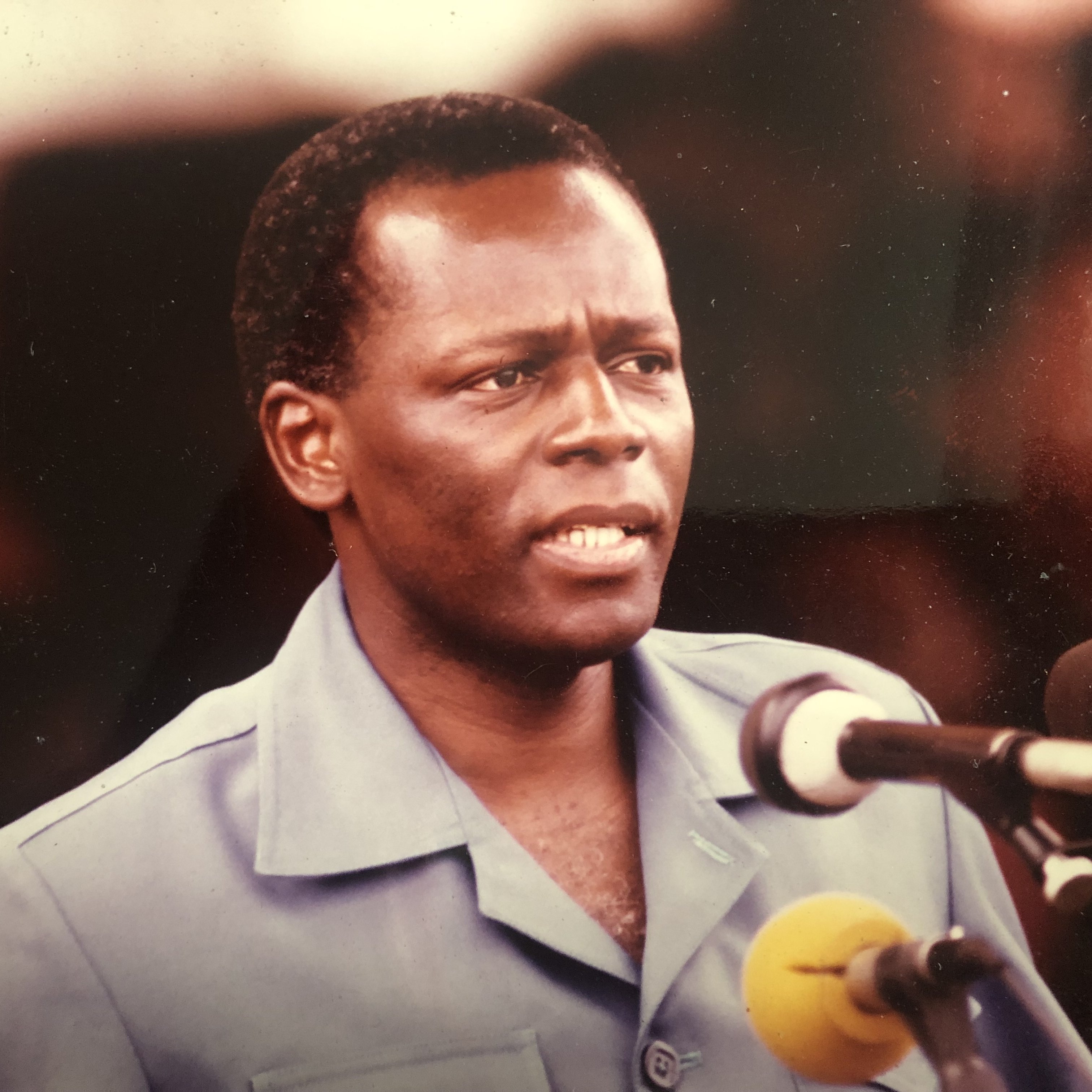 L'image montre un homme en train de prononcer un discours devant un microphone. Il porte une chemise de couleur bleue et semble sérieux et engagé dans ses propos. Son visage exprime une certaine détermination, et il se tient droit. Il y a plusieurs microphones devant lui, et un public peut être présent en arrière-plan, bien que non visible sur cette image. L'atmosphère donne l'impression d'un moment important lors d'un discours ou d'une déclaration publique.