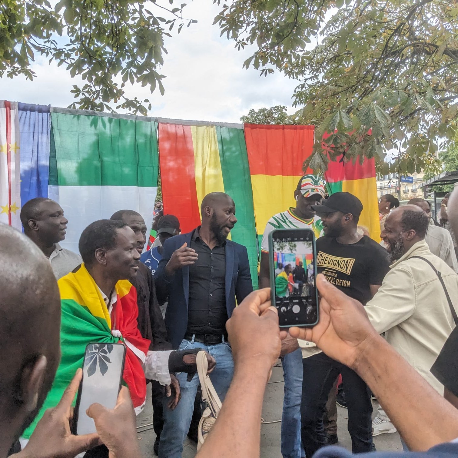L'image montre un groupe de personnes réunies autour d'un fond coloré constitué de drapeaux. Les drapeaux affichés incluent ceux du Cameroun, de l'Union européenne, du Mali et de la République de Guinée. Des hommes portent des vêtements variés, certains drapés dans des drapeaux, ce qui suggère un événement festif ou culturel. Ils semblent en discussion animée, souriant et échangeant des plaisanteries. Au premier plan, un participant tient un téléphone portable, probablement pour capturer le moment. L'atmosphère semble conviviale et pleine d'énergie.