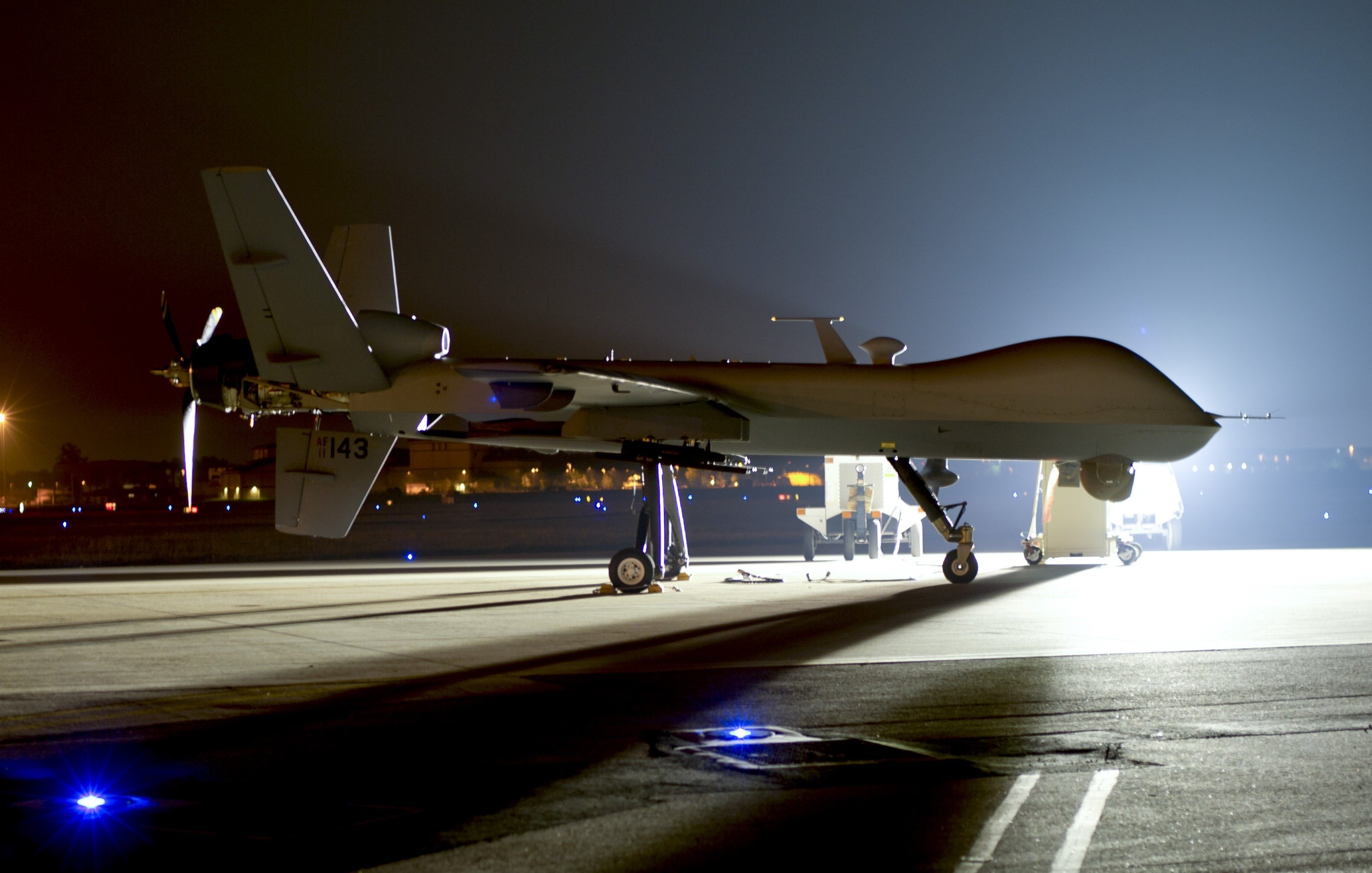 L'image montre un drone de surveillance, typiquement de type militaire, stationné sur une piste d'atterrissage. La scène est nocturne, éclairée principalement par des lumières artificielles qui révèlent les contours du drone. Le drone a une forme allongée et est équipé d'une hélice à l'arrière. Il repose sur un châssis avec des supports, et il y a des lumières au sol qui peuvent délimiter la zone ou indiquer l'emplacement. L'ambiance est sérieuse et technologique, évoquant des missions de surveillance ou de reconnaissance.