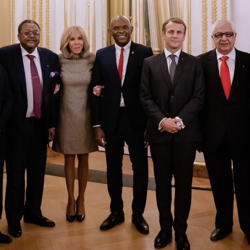 L'image montre un groupe de sept personnes se tenant debout dans une salle élégante, avec des murs clairs et des rideaux drapés. Au centre, une femme souriante porte une robe brillante et se tient entourée de six hommes en costume. Chacun d'eux a un regard chaleureux et engageant. Les hommes varient en teinte de peau et en style vestimentaire, certains portant des cravates, d'autres des vêtements traditionnels. L'ambiance de l'image dégage une atmosphère professionnelle et amicale, suggérant un moment de convivialité ou une rencontre importante.