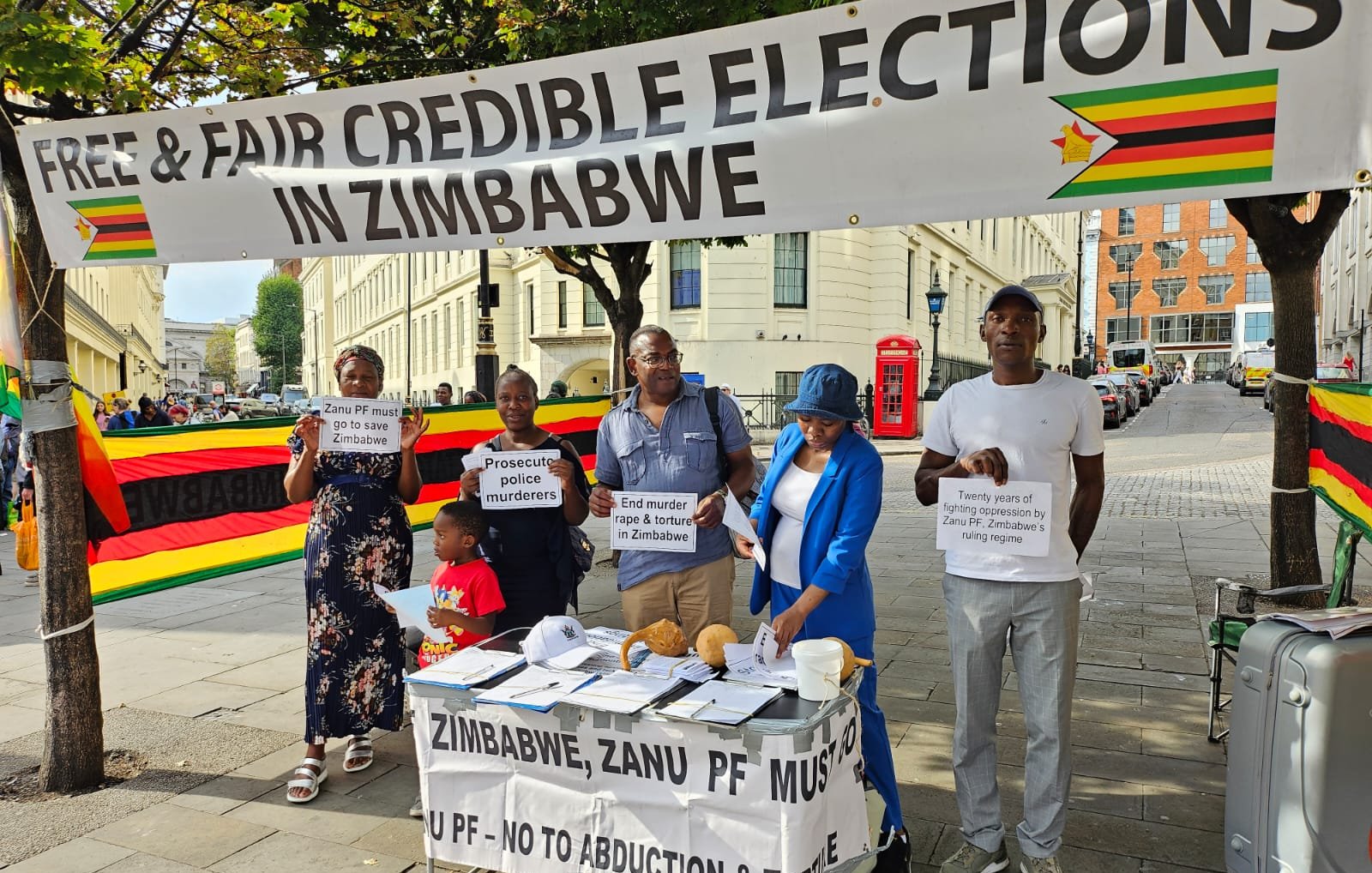 L'image montre un groupe de personnes rassemblées sous une grande banderole qui proclame "Élections libres et équitables au Zimbabwe". Cette banderole est ornée des couleurs du drapeau zimbabwéen, avec du rouge, du jaune, du vert et du noir. Les manifestants tiennent des panneaux avec des messages portant sur la nécessité de respecter les droits humains et d'organiser des élections justes au Zimbabwe. Sur la table devant eux, il y a des brochures et des documents qui semblent orientés vers la sensibilisation et l'information sur la situation politique. L'environnement est urbain, avec des bâtiments en arrière-plan et des passants qui s'intéressent à la manifestation.