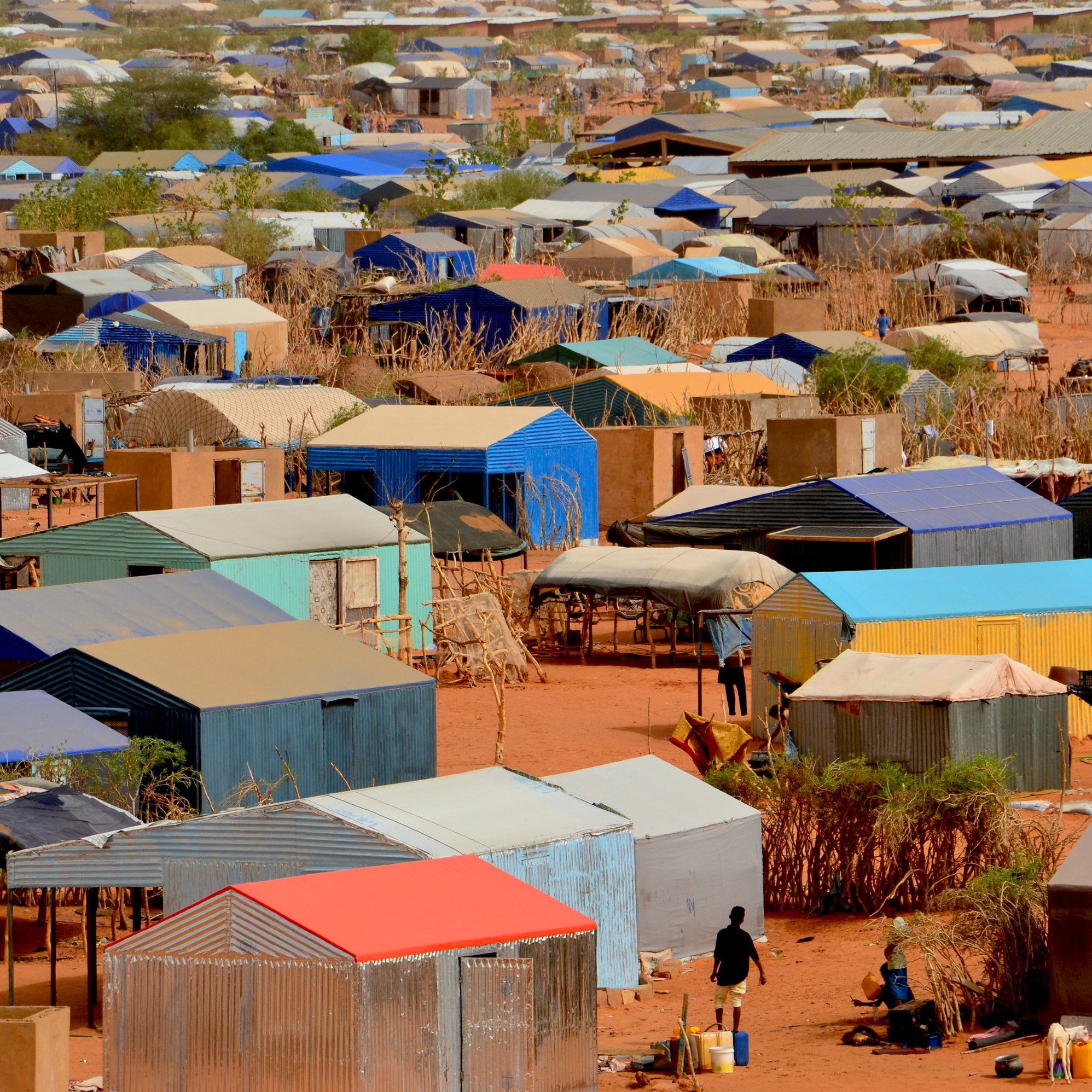 L'image montre un vaste camp de tentes et de logements temporaires disposés de manière dense sur un terrain désertique. Les structures sont recouvertes de toits de différentes couleurs, principalement des teintes de bleu, de rouge et de gris. Le sol est sablonneux, et quelques arbres ou buissons apparaissent ici et là, apportant un peu de verdure. En bas de l'image, on peut apercevoir des personnes se déplaçant entre les tentes, évoquant une activité quotidienne dans ce milieu. L'ensemble dégage une impression d'organisation dans la diversité des abris, tandis que le paysage aride souligne les conditions de vie dans ce camp.