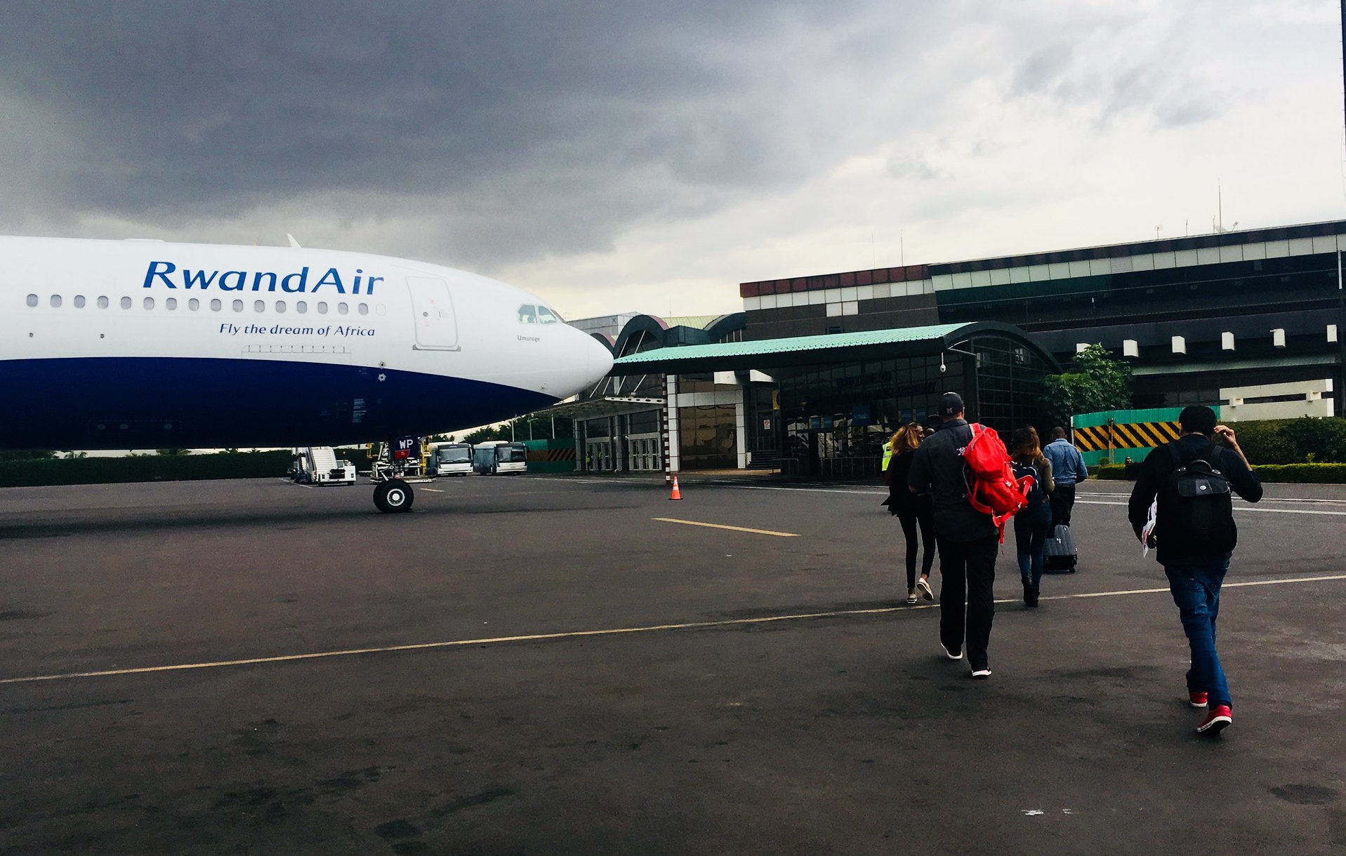 L'image montre un groupe de personnes marchant vers un avion de la compagnie RwandAir, qui est garé sur une aire d'aéroport. L'avion, avec une livrée bleue et blanche, est imposant avec un nez bien visible. Le ciel est nuageux, laissant présager une ambiance légèrement sombre. À l'arrière-plan, on voit un bâtiment d'aéroport avec une architecture moderne. Les voyageurs, portant des sacs à dos et d'autres bagages, avancent vers l'avion, créant un sentiment de départ et d'aventure. Le sol est en béton, et il y a des cônes de signalisation dispersés sur le terrain.