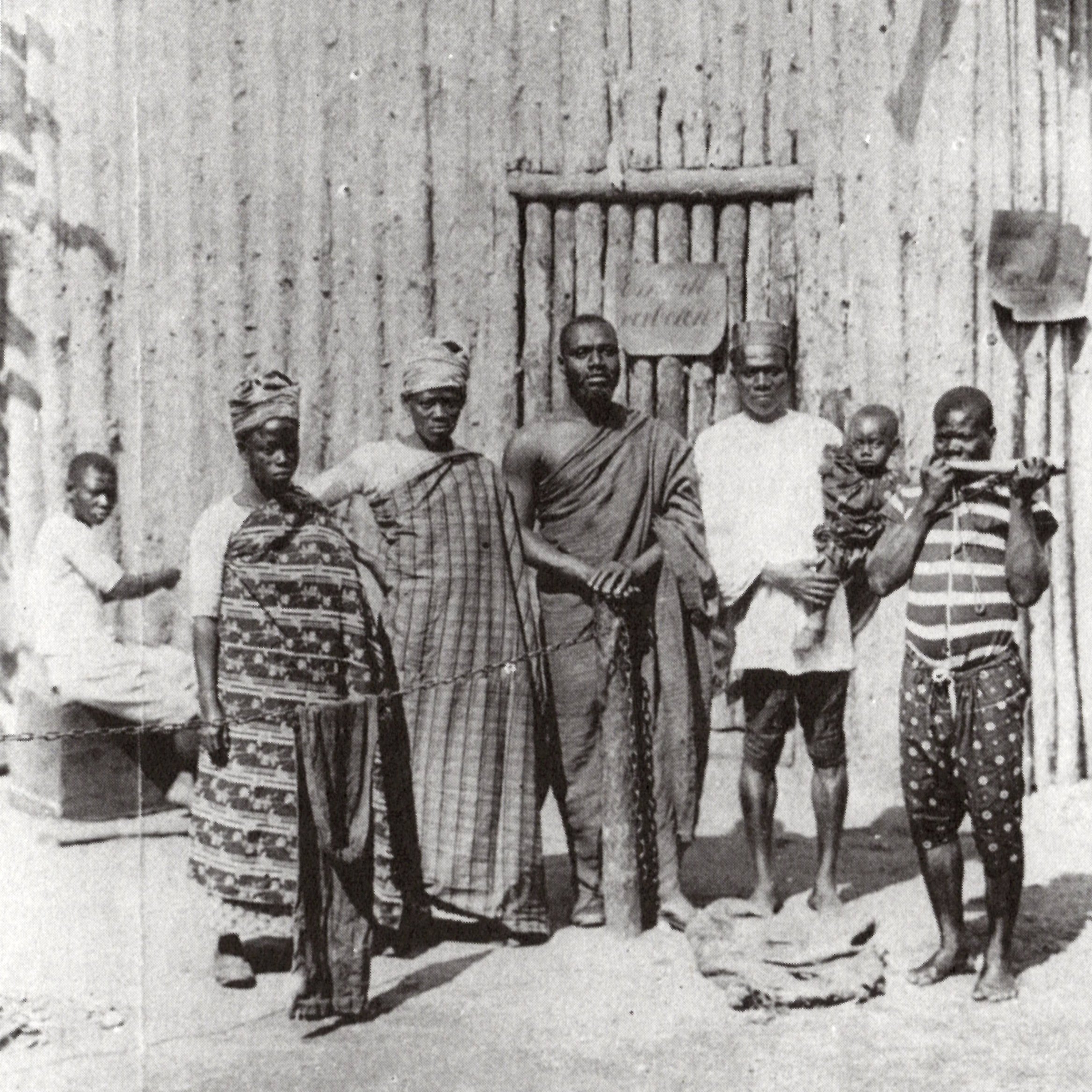 L'image montre un groupe de six personnes se tenant debout devant une structure en bois, qui semble être une maison ou un abri, avec un décor rustique. Au fond, on distingue un homme assis qui fait face à la scène principale. Les membres du groupe portent des vêtements traditionnels typiques, avec des motifs variés, affichant une richesse culturelle. L'un d'eux tient un enfant, tandis qu'un autre est équipé d'un bâton, suggérant une certaine autorité ou dignité. L'atmosphère de l'image dégage un sentiment de communauté et de convivialité.