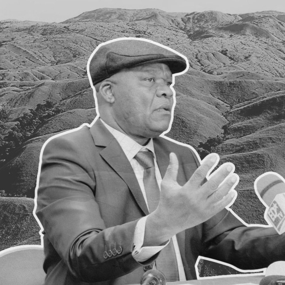 L'image montre un homme en costume, avec un chapeau, en train de parler lors d'une conférence ou d'une interview. Il est assis devant un microphone et semble gesticuler pour appuyer ses propos. En arrière-plan, on aperçoit un paysage de collines ondulantes, évoquant une nature sauvage et montagneuse, donnant une impression de profondeur et de calme. L'ensemble de l'image est en noir et blanc, ce qui renforce le sérieux de la scène.