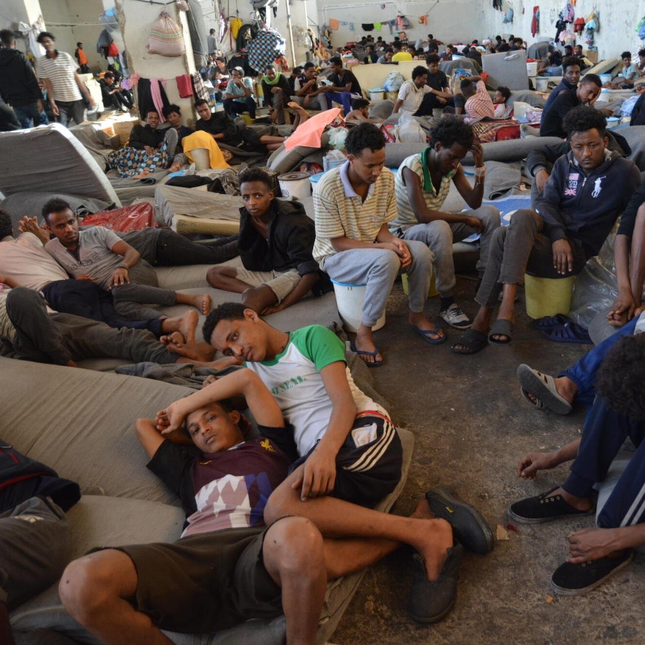 L'image montre un grand espace intérieur, probablement une salle ou un centre d'accueil, où de nombreuses personnes sont rassemblées. Le sol est recouvert de matelas ou de couvertures, et les gens sont assis ou allongés, souvent en petits groupes ou seuls. Les visages montrent une variété d'émotions, allant de la fatigue à l'attente. L'atmosphère semble chargée, et l'agencement des personnes donne une idée d'une situation difficile. Les murs sont nus, et il y a des effets personnels éparpillés ici et là. L'environnement est sombre et témoigne d'un manque de confort.