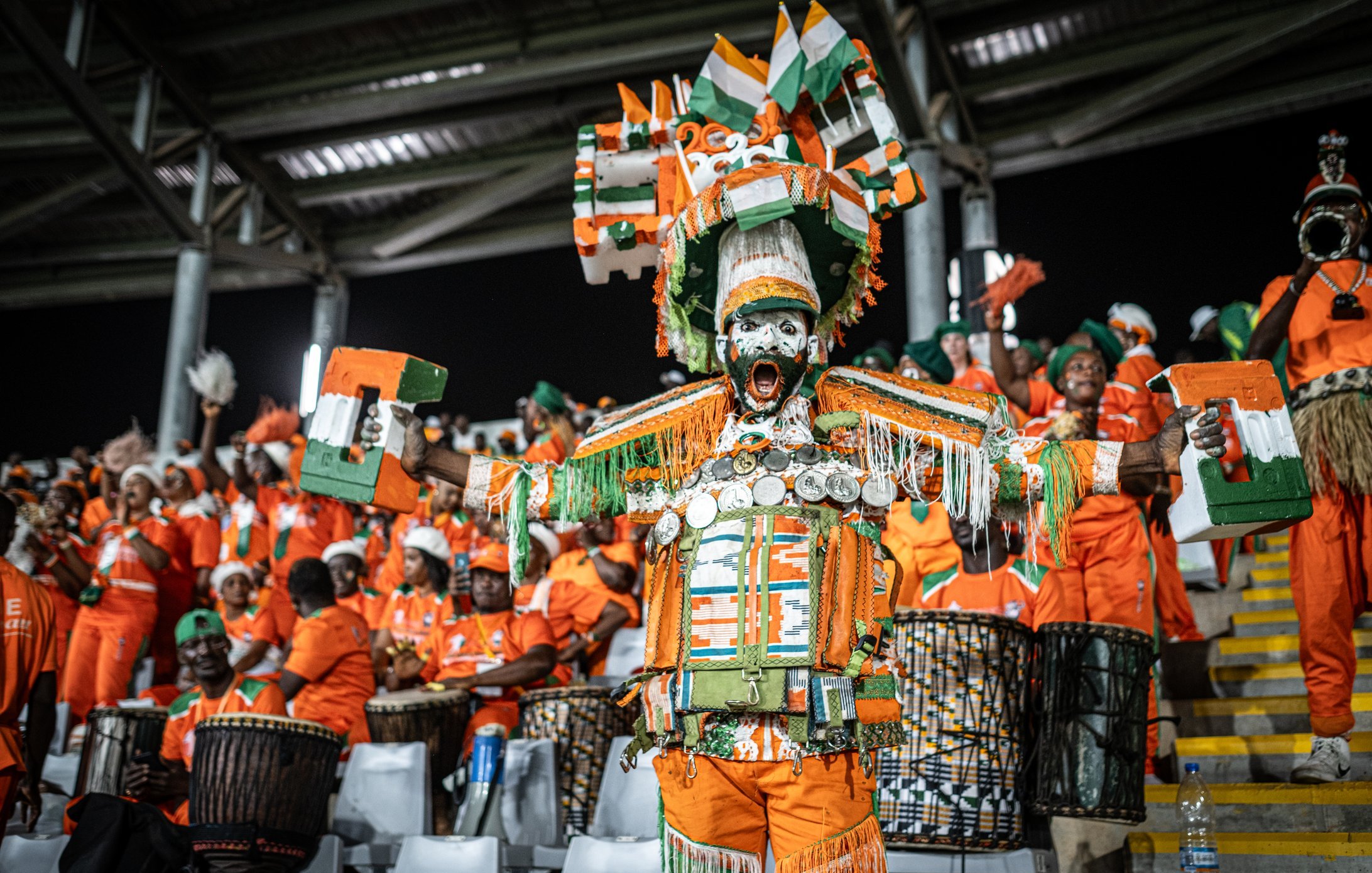 L'image montre une scène vibrante d'un événement festif, probablement un match de football. Au premier plan, un homme se distingue par son costume coloré composé de tissus orange et de nombreux accessoires décoratifs, dont des coquillages et des éléments en métal qui brillent à la lumière. Il est entouré de groupes de personnes, également en orange, qui jouent des tambours et sont vêtus de vêtements similaires. Tous semblent animés et enthousiastes, contribuant à une atmosphère de fête collective, ponctuée de chants et de danses. Les visages sont exprimés par des sourires et de la joie, avec des éléments traditionnels mettant en avant la culture locale.