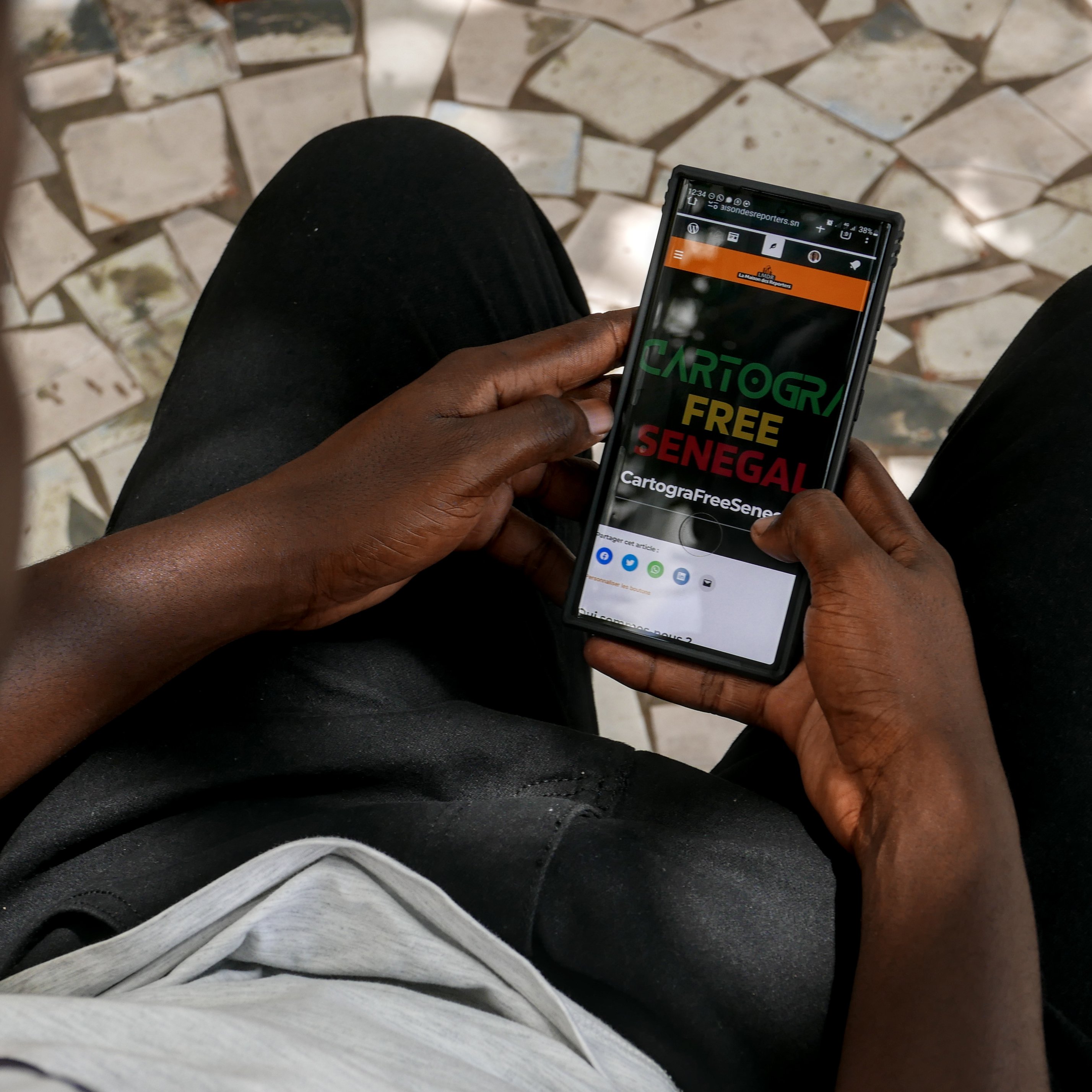 Sur cette image, on voit une personne assise, tenant un smartphone dans ses mains. La personne semble attentive à l'écran, qui affiche un site ou une application avec les mots "CARTOGR" en gros caractères, suivi de "FREE SENEGAL". Le fond est constitué de carreaux de mosaïque, tandis que la personne porte un t-shirt clair et un pantalon noir. L'arrière-plan et la position des mains indiquent une ambiance calme et concentrée.