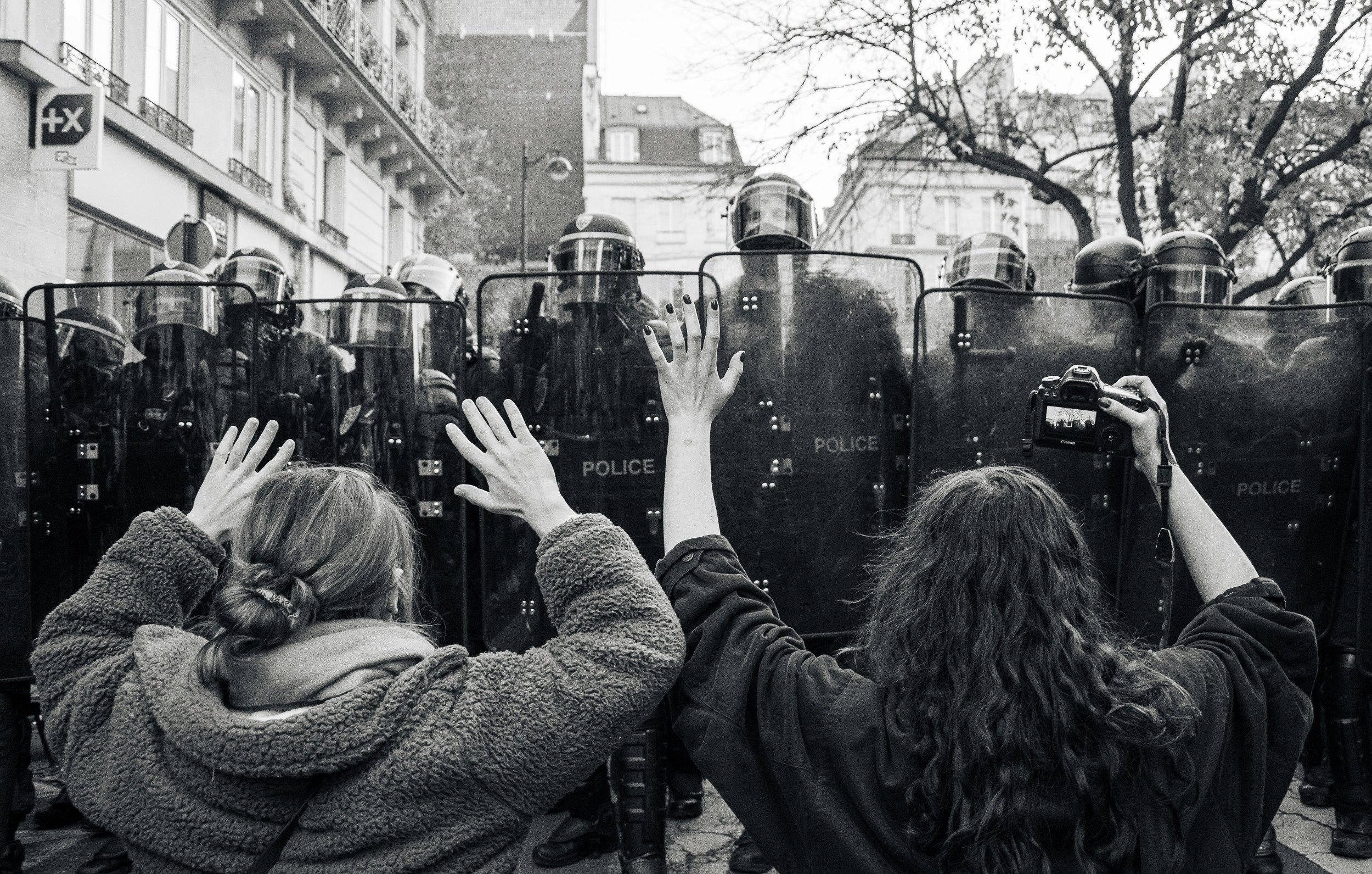 L'image montre une scène de manifestation en noir et blanc. À l'avant-plan, deux personnes se tiennent face à des policiers en tenue anti-émeute. Les manifestants, une femme brune avec les cheveux longs et une autre avec des cheveux bouclés, lèvent les bras en signe de protestation ou de désescalade. Les policiers sont rangés en formation derrière des boucliers, créant une barrière. L'environnement urbain est visible en arrière-plan, avec des bâtiments peut-être typiques de la ville. L'atmosphère est tendue, reflétant un moment de confrontation pacifique mais chargée d'émotion.