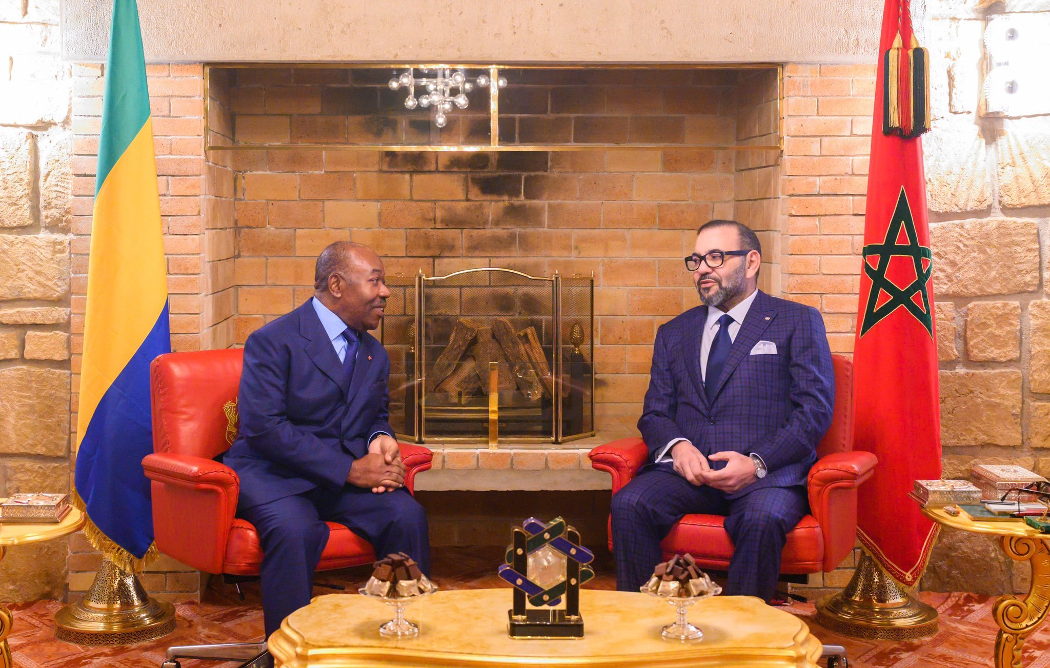 L'image montre deux hommes assis face à face dans un salon chaleureux et élégant. À gauche, un homme en costume bleu, avec une cravate, est légèrement incliné vers l'avant, écoutant attentivement. À droite, un autre homme porte un costume à carreaux bleu foncé et une cravate assortie, affichant une expression sérieuse tout en participant à la conversation. Derrière eux, on peut voir un mur en briques claires et une cheminée décorative. Il y a également des éléments de décoration comme des fauteuils rouges et un tableau avec un drapeau marocain sur le côté droit. Une lumière douce illumine la pièce, créant une atmosphère conviviale.