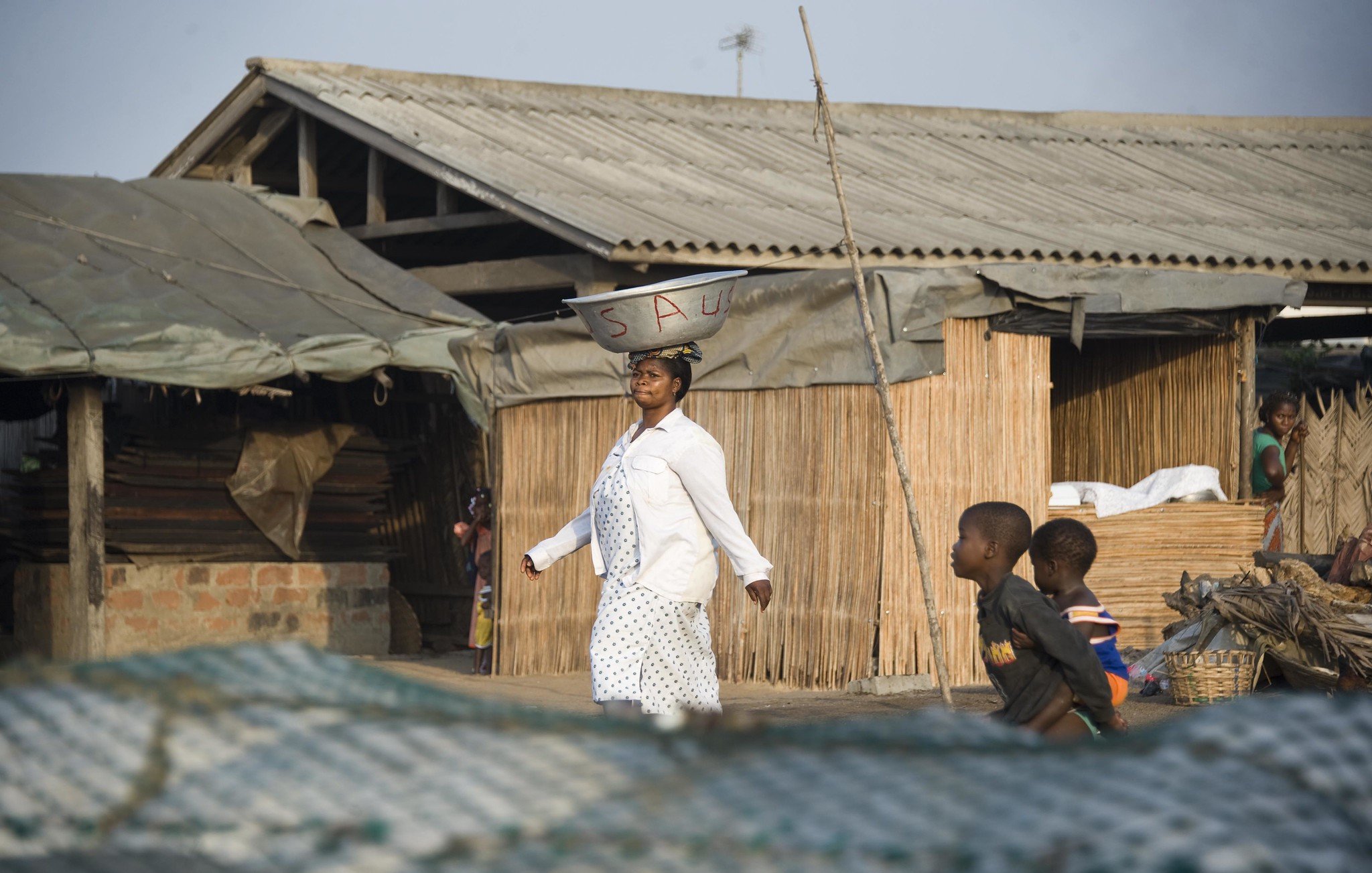 Dans cette scène vibrante, une femme porte une grande bassine sur sa tête, symbole de son travail quotidien. Elle avance avec assurance sur un chemin, tout en étant entourée de maisons à architecture simple, en bois et en bambou. Le ciel est clair, laissant présager une belle journée ensoleillée. À proximité, des enfants jouent ensemble, ajoutant une atmosphère de vie et de dynamisme à l'environnement. Les ombres et les textures des matériaux environnants se mêlent, créant un paysage riche et vivant. L'ensemble dégage une ambiance d'activité et de communauté.