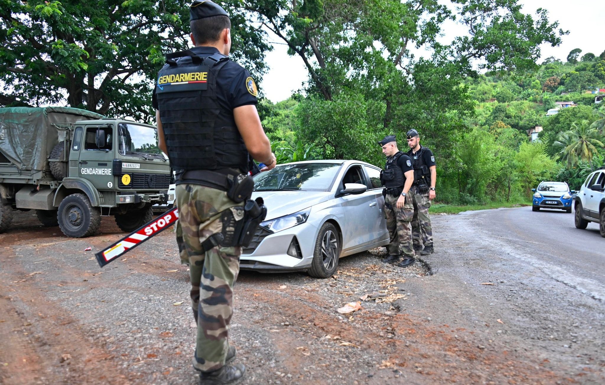 L'image montre un contrôle routier en plein air. Au premier plan, un gendarme en uniforme, portant un gilet par balles noir avec l'inscription "Gendarmerie", observe une voiture argentée qui est arrêtée sur le côté de la route. La voiture a un panneau "STOP" visible. Deux autres gendarmes, habillés de manière similaire, se tiennent près de la voiture, dialoguant probablement avec le conducteur. En arrière-plan, on aperçoit une camionnette militaire de la gendarmerie et des arbres verdoyants qui donnent une ambiance naturelle à la scène. Le sol est en terre, avec beaucoup de verdure autour, ce qui indique que le lieu est rural. L'atmosphère semble sérieuse, reflet du contrôle de sécurité en cours.