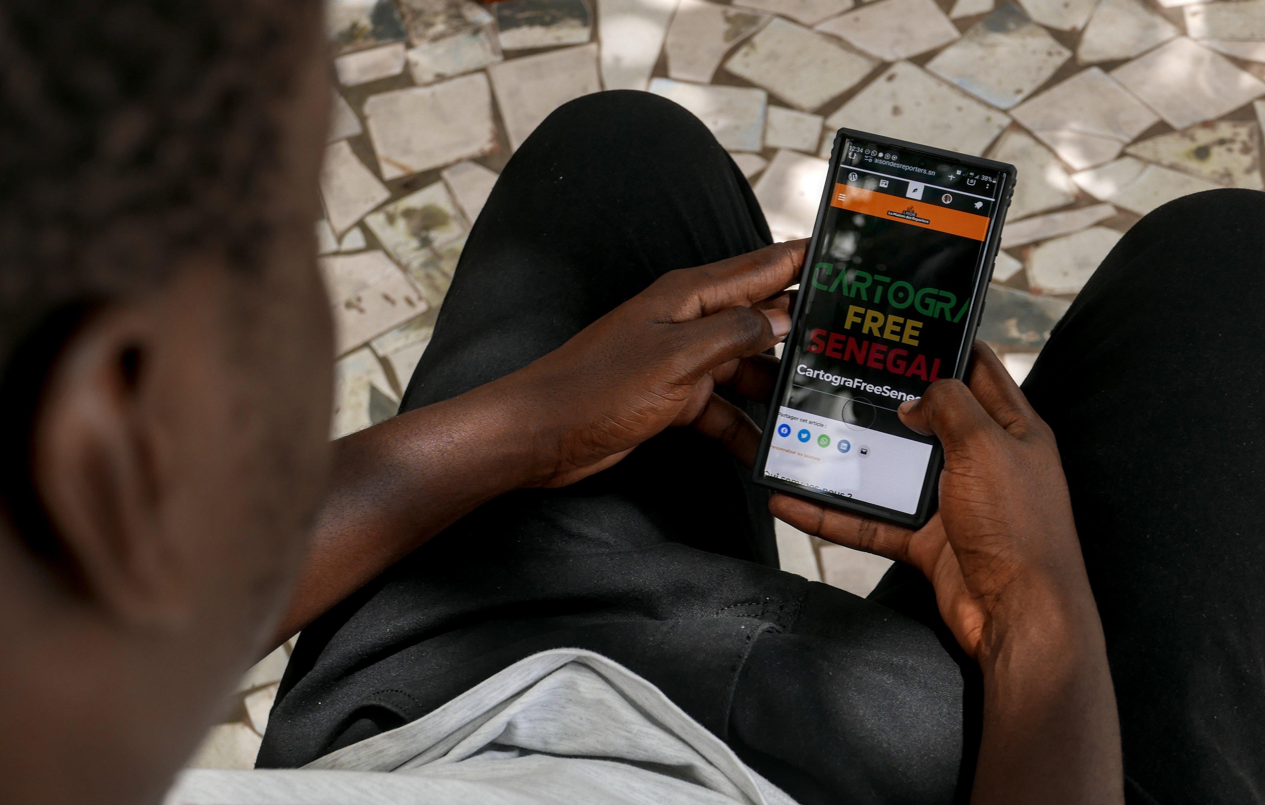 Sur cette image, on voit une personne assise, tenant un smartphone dans ses mains. La personne semble attentive à l'écran, qui affiche un site ou une application avec les mots "CARTOGR" en gros caractères, suivi de "FREE SENEGAL". Le fond est constitué de carreaux de mosaïque, tandis que la personne porte un t-shirt clair et un pantalon noir. L'arrière-plan et la position des mains indiquent une ambiance calme et concentrée.