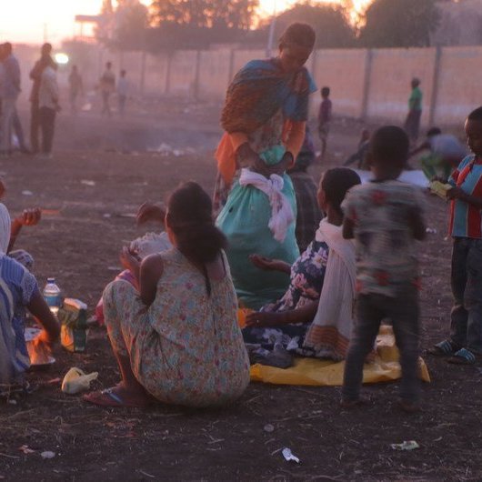 L'image montre une scène de vie en plein air, probablement à la tombée de la nuit. Au premier plan, plusieurs femmes et enfants sont assis sur le sol, entourés de quelques objets personnels et de restes de nourriture. Les femmes, habillées de vêtements colorés, semblent préparer un repas autour d'un feu. En arrière-plan, la lumière du crépuscule illumine légèrement la scène, et de nombreuses personnes peuvent être aperçues se déplaçant dans un environnement poussiéreux. L'atmosphère dégage à la fois une certaine convivialité et une sensation de précarité.