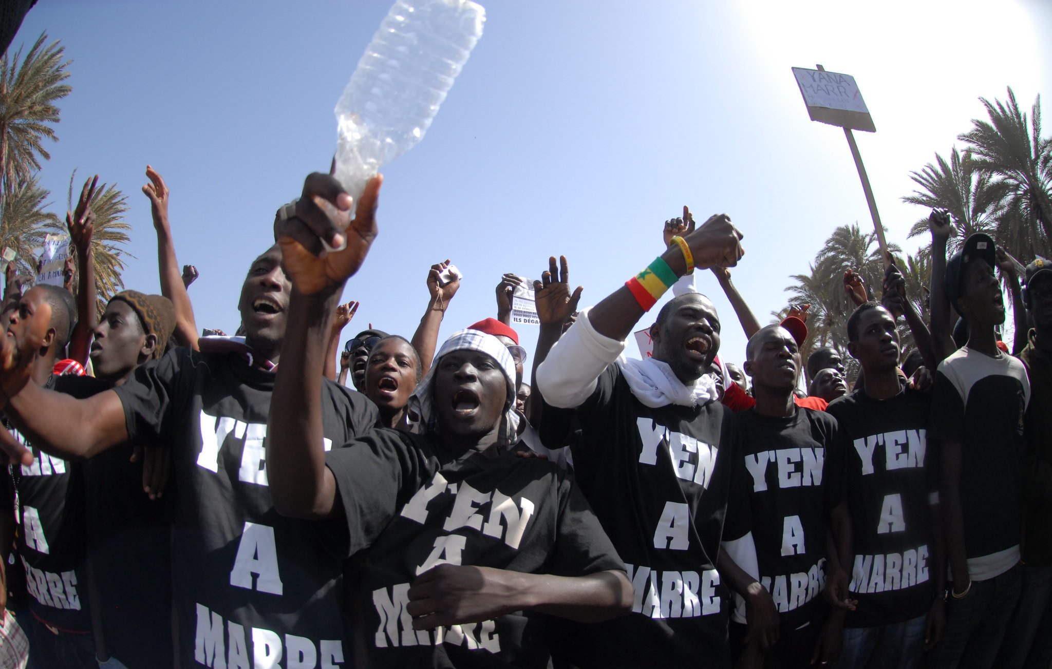 L'image montre un groupe de personnes rassemblées pour exprimer leur mécontentement, probablement lors d'une manifestation. Les membres du groupe portent des t-shirts noirs avec les mots "Y'en a marre" inscrits en lettres blanches, ce qui signifie qu'ils en ont assez d'une situation particulière. On peut les voir lever les bras, certains brandissant des bouteilles en plastique, et afficher des visages déterminés. En arrière-plan, on aperçoit des palmiers et un ciel bleu, indiquant une ambiance ensoleillée. L'énergie de la scène est palpable, avec des cris et une forte intensité émotionnelle.