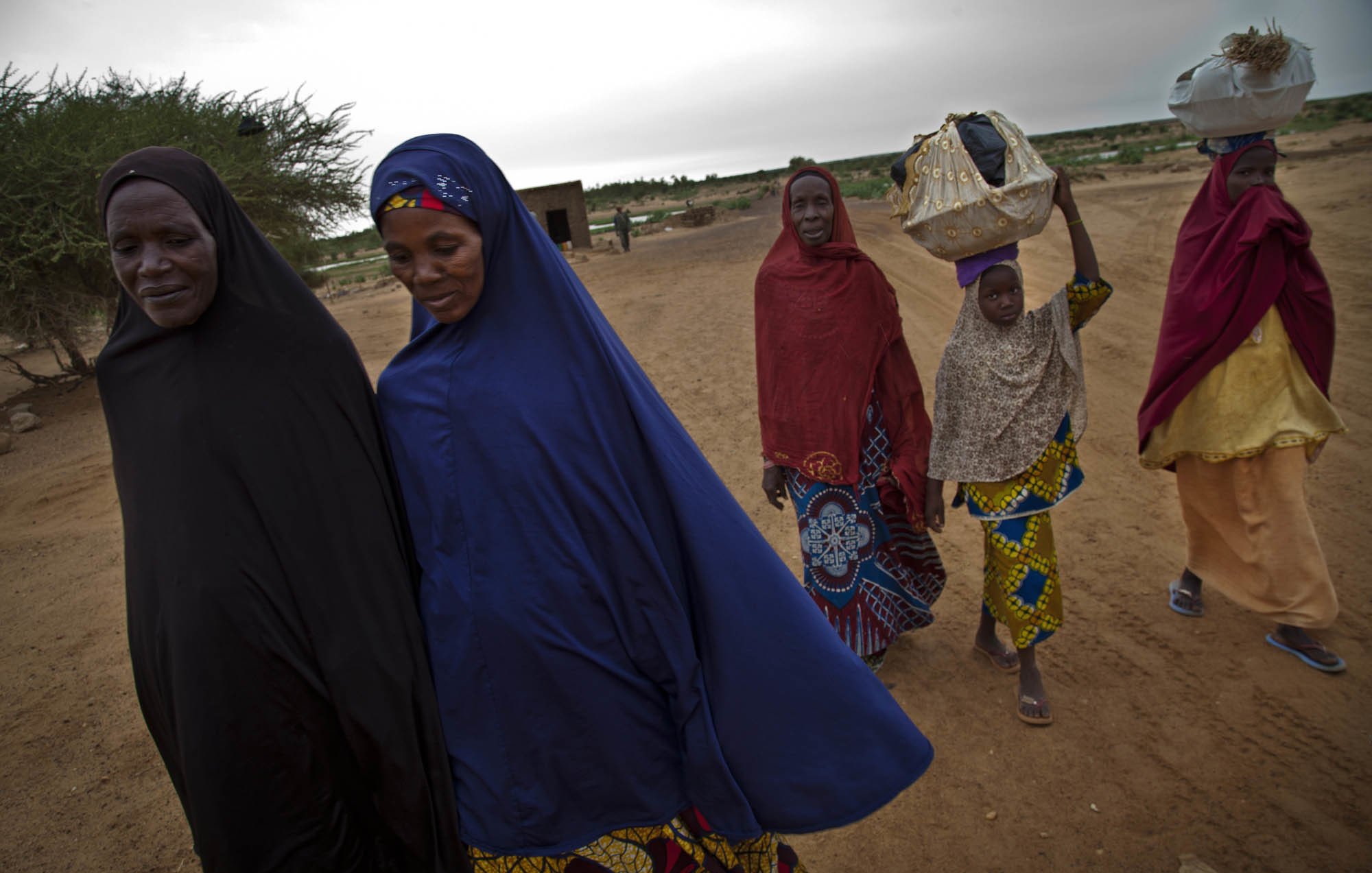 L'image représente un groupe de cinq femmes marchant sur un chemin de terre. Elles portent des vêtements traditionnels colorés, avec des motifs variés. Certaines femmes ont un voile couvrant leur tête, et une jeune fille à l'arrière transporte un objet sur sa tête, probablement un panier. Le paysage environnant est sec et désertique, avec quelques arbustes épars en arrière-plan, et le ciel est couvert, suggérant une ambiance calme et sereine. L'ensemble transmet une impression de solidarité et de culture, avec un accent sur les traditions vestimentaires.