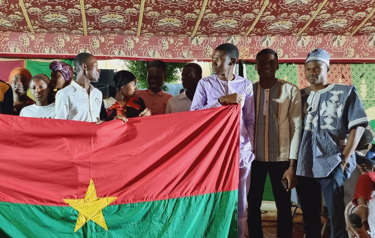 The image depicts a group of young people gathered together, holding a large flag of Burkina Faso, which features a red field with a green stripe at the bottom and a yellow star in the center. The setting appears to be festive, possibly during a cultural or community celebration, with decorative elements in the background. The individuals are wearing a mix of traditional and modern clothing, smiling and posing together, suggesting a sense of unity and pride in their national identity.