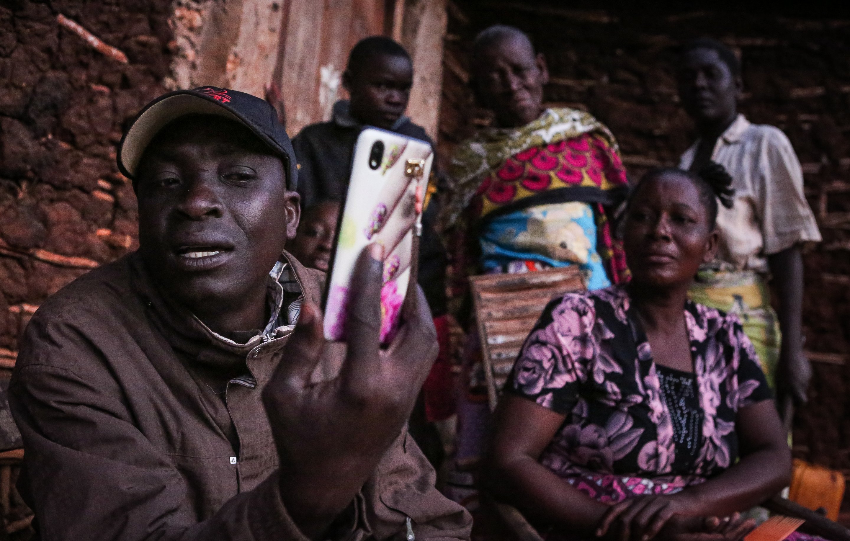 Cette image présente un groupe de personnes rassemblées à l'extérieur d'une maison. Au premier plan, un homme tient un téléphone portable et semble communiquer ou montrer quelque chose à ses interlocuteurs. Il porte une casquette et un manteau. À l'arrière-plan, plusieurs femmes regardent avec attention. Elles sont vêtues de vêtements colorés et affichent des expressions d'intérêt ou de curiosité. L'environnement est simple, avec des murs en terre crue et un éclairage qui suggère qu'il pourrait être tard dans la journée. L'atmosphère semble conviviale et axée sur la communication.