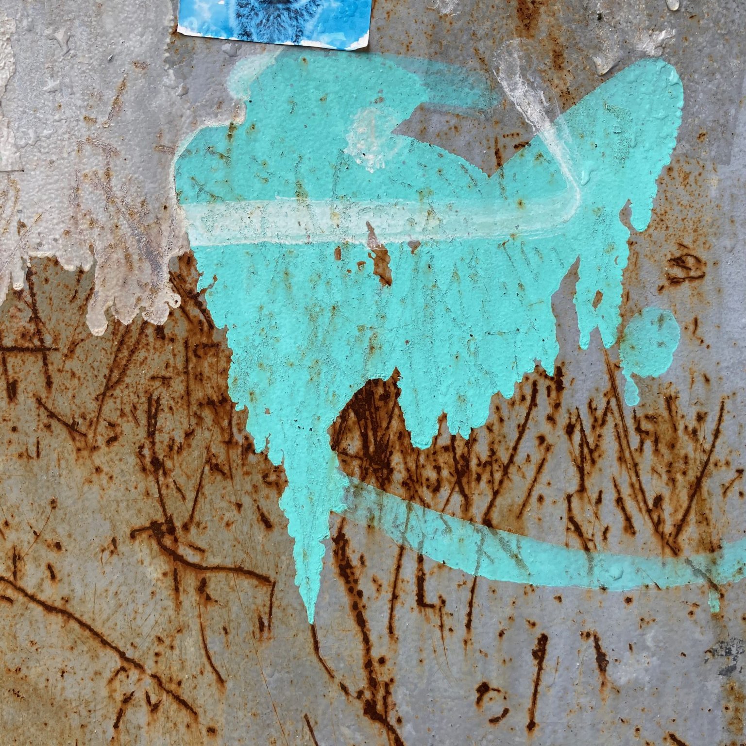 L'image présente une surface en métal, probablement rouillée, avec des marques et des textures variées. On y voit une tache de couleur turquoise qui semble avoir été projetée. Cette tache a une forme fluide et irrégulière, évoquant une sorte de mouvement ou de dynamisme. À proximité, on peut distinguer des traces brunes, résultant de la rouille, qui ajoutent un contraste marqué à la couleur vive. Le fond est texturé, avec des stries et des imperfections, suggérant l'âge et l'usure du matériau. L'ensemble crée une atmosphère à la fois urbaine et artistique.
