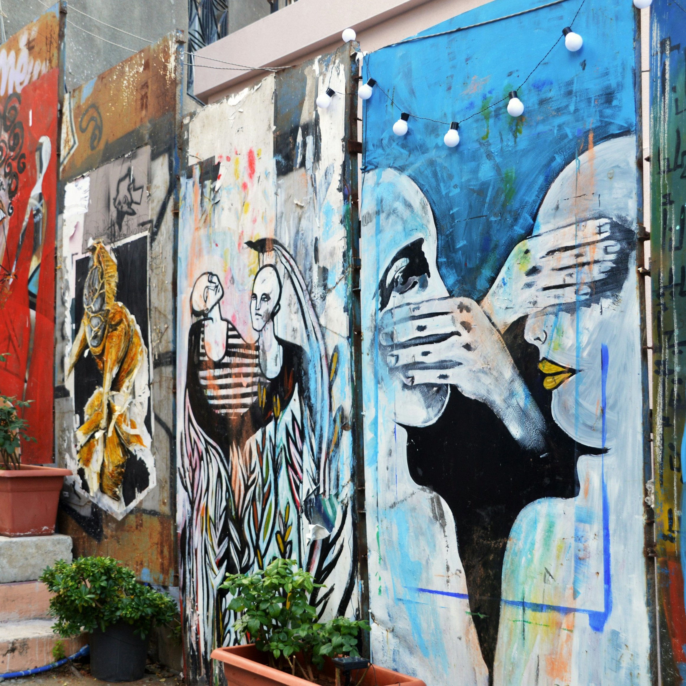 L'image présente un mur de graffitis colorés qui ornent une ruelle. On peut distinguer plusieurs œuvres d'art, chacune ayant son propre style. À gauche, une figure regardante avec des détails réalistes, suivie d'illustrations plus abstraites et expressives au centre et à droite, représentant des visages et des mains en mouvement. Les couleurs dominantes incluent des nuances vives de bleu, d'orange et de noir. Des pots de fleurs, avec des plantes vertes, ajoutent une touche de nature à l'environnement urbain. Au-dessus des œuvres, des guirlandes lumineuses créent une ambiance chaleureuse. Les escaliers en pierre accompagnent le mur, suggérant une connexion avec l'espace environnant. L'ensemble dégage une atmosphère artistique et dynamique, reflet d'un lieu vivant et créatif.