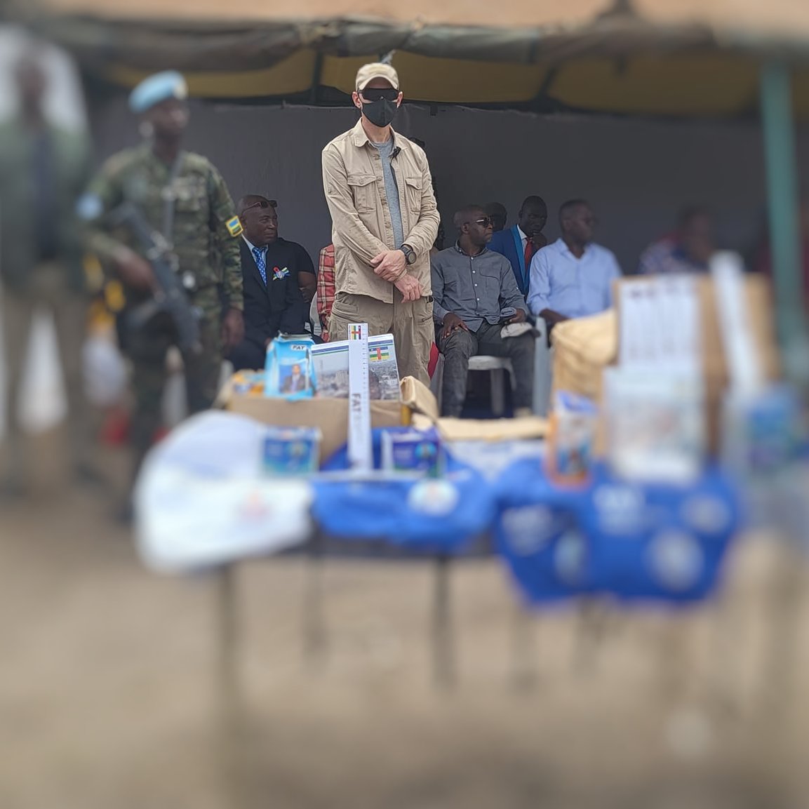 L'image montre un événement en plein air, avec une tente sur le côté. Au centre, on voit une table couverte d'objets et de brochures, probablement liés à une présentation ou un programme. À l'arrière-plan, plusieurs personnes sont assises, y compris des enfants et des adultes. Un homme se tient debout, portant une casquette et un masque, semblant être un intervenant ou une personne d'autorité. La scène reflète un moment de rassemblement communautaire, chargé d'une atmosphère d'échange et d'information.