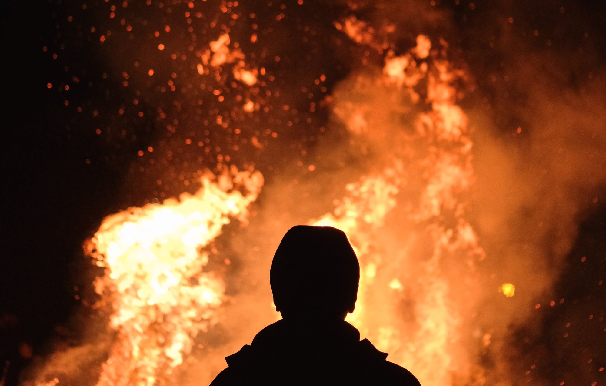 L'image montre une scène nocturne où une personne se tient de dos, face à un grand feu. Les flammes dansent avec éclat, projetant une lumière vive et chaleureuse dans l'obscurité environnante. Des étincelles s'élèvent en spirales, créant une ambiance à la fois magique et intense. La silhouette de la personne est sombre, contrastant avec les couleurs vives de l'incendie, ce qui permet de ressentir la chaleur émanant du feu et l'atmosphère captivante qui l'entoure.