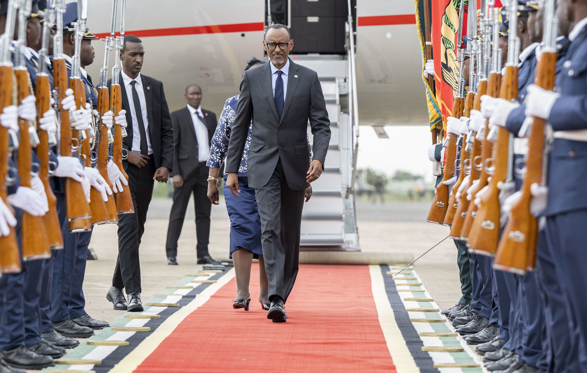 L'image montre un moment officiel sur un tapis rouge, où un homme en costume, probablement un dignitaire, marche avec assurance. Il est entouré de garde d'honneur en tenue formelle, tenant des fusils. À l'arrière-plan, on aperçoit une grande échelle d'avion et d'autres personnes en costume. L'atmosphère dégage un sentiment de cérémonie et de respect, soulignée par la disposition soignée des militaires et le faste du décor.