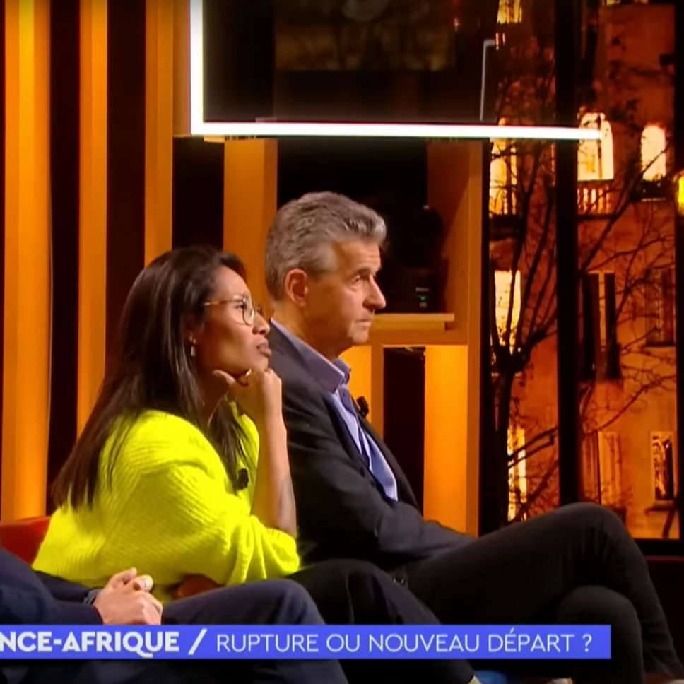 L'image montre un plateau de télévision lors d'une émission intitulée "France-Afrique : Rupture ou nouveau départ ?". Trois personnes sont assises sur un canapé, éclairées par des lumières chaudes. À gauche, un homme avec des lunettes et des cheveux grisonnants porte un pull sombre. Au centre, une femme, habillée d'un pull lumineux, semble attentive et réfléchie. À droite, un homme aux cheveux courts et argentés affiche une expression sérieuse. En arrière-plan, on peut apercevoir des fenêtres illuminées qui donnent une ambiance chaleureuse à l'ensemble. Le décor est moderne, avec des éléments graphiques en arrière-plan qui ajoutent à l'esthétique de l'émission.