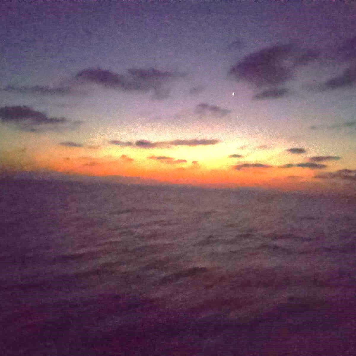 The image shows a serene seascape during twilight. The sky is transitioning from daylight to night, displaying a gradient of colors ranging from deep blue to orange near the horizon. Clouds are scattered across the sky, adding to the atmospheric depth. A single bright star is visible in the upper right portion of the image, contributing to the tranquil mood of the scene. The calm ocean reflects these colors, creating a peaceful and beautiful setting.