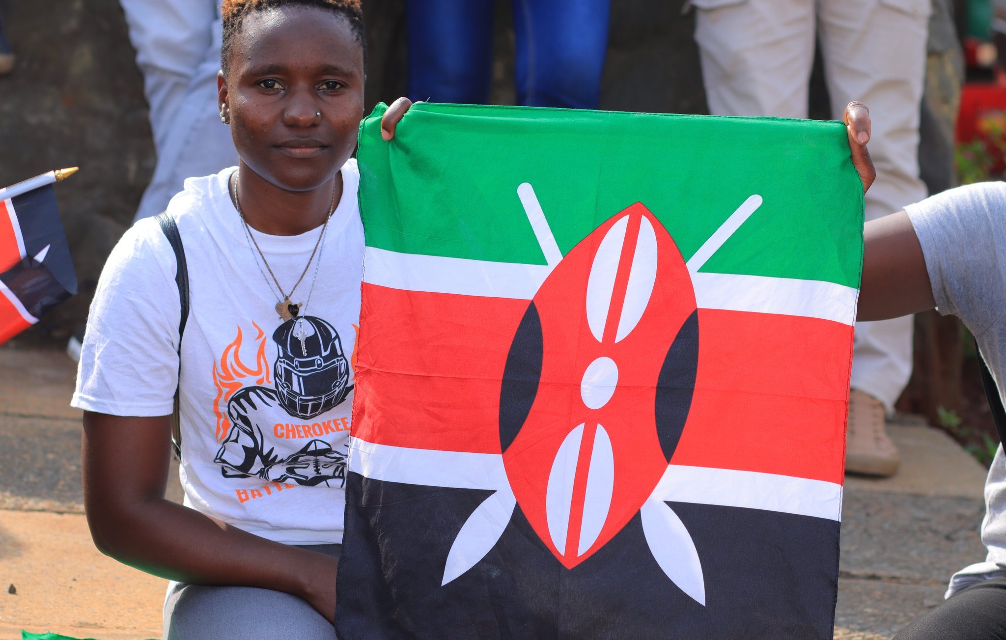 Dans cette image, une jeune femme sourit tout en tenant une grande drapeau du Kenya. Le drapeau est composé de bandes horizontales de couleur noire, rouge et verte, avec un motif central représentant un écu et des flèches. La femme porte un t-shirt blanc orné d'un motif sportif, avec un choker en pendentif autour du cou. En arrière-plan, on peut apercevoir d'autres personnes, certaines tenant également des drapeaux, créant une ambiance festive et patriotique.