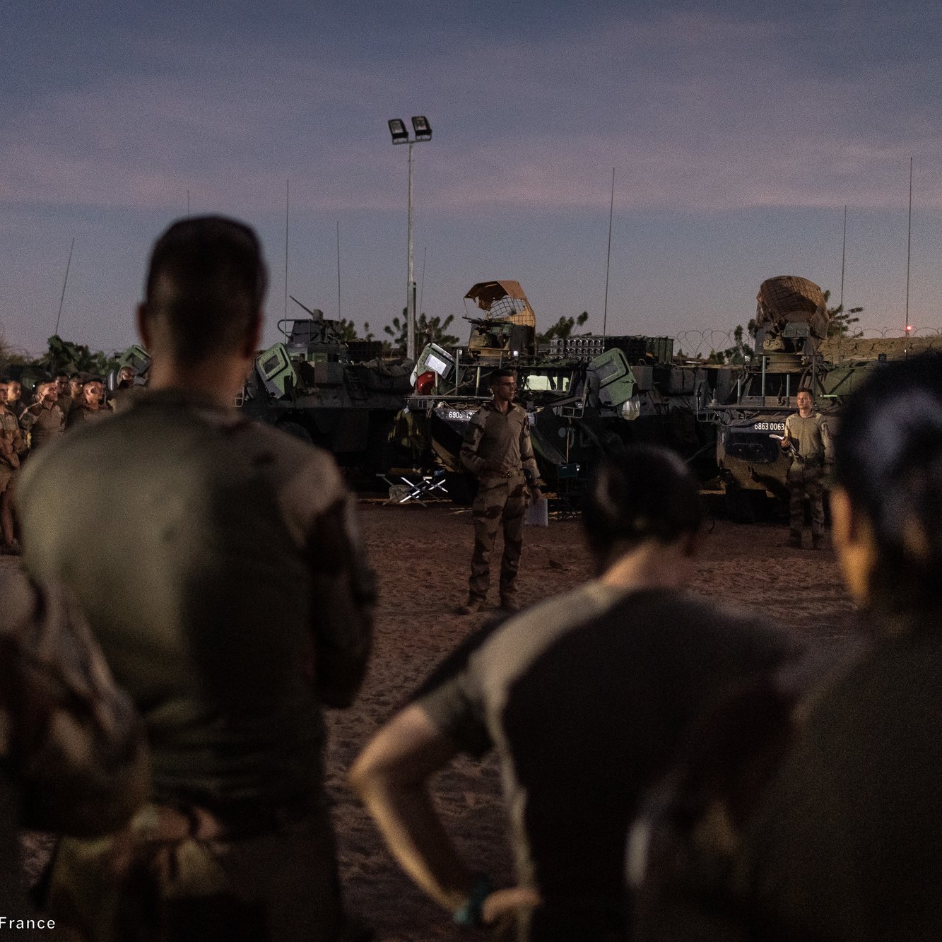 L'image montre un groupe de soldats rassemblés, attentifs, dans un environnement désertique au crépuscule. Ils sont partiellement de dos, ce qui met en avant leur tenue militaire. À l'arrière-plan, plusieurs véhicules militaires sont visibles, équipés d'armements. Le ciel est teinté de nuances douces, marquant la transition entre le jour et la nuit. L'atmosphère semble concentrée, suggérant une préparation ou un briefing important pour les soldats sur le terrain.