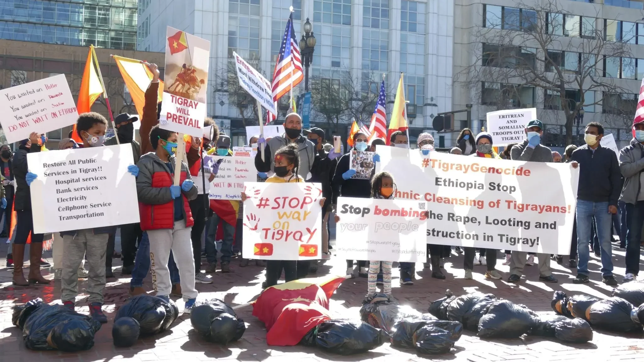 L'image montre une manifestation animée dans un espace urbain. Des personnes se tiennent debout avec des pancartes et des drapeaux, exprimant leur soutien pour la cause du Tigré. Les pancartes affichent des slogans tels que "Tigray Genocide" et "Stop bombing your people", dénonçant la violence et la situation humanitaire dans cette région d'Éthiopie. Au sol, on aperçoit des sacs noirs, symbolisant des victimes ou une tragédie. L'atmosphère est chargée d'émotion, avec une foule engagée, destinée à attirer l'attention sur cette crise.