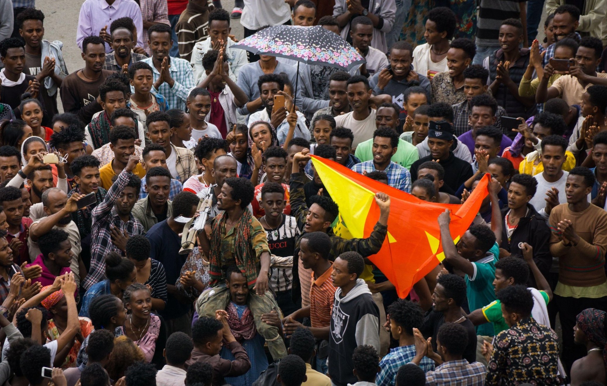 L'image représente une scène vibrante et animée d'une foule rassemblée ensemble. Au premier plan, on peut voir un groupe dense de personnes, principalement des jeunes hommes, exprimant leur énergie et leur enthousiasme. Certains d'entre eux lèvent les bras, d'autres applaudissent ou tiennent des drapeaux aux couleurs vives, notamment le rouge et le jaune. Au centre, une personne semble parler ou chanter, attirant l'attention de la foule. Les visages expriment des émotions variées telles que la joie, la passion et la détermination. Par temps peut-être nuageux, un parapluie coloré est en arrière-plan, ajoutant une note de couleur à la scène. L'ambiance générale est celle d'un rassemblement convivial, où les participants partagent un moment fort ensemble.