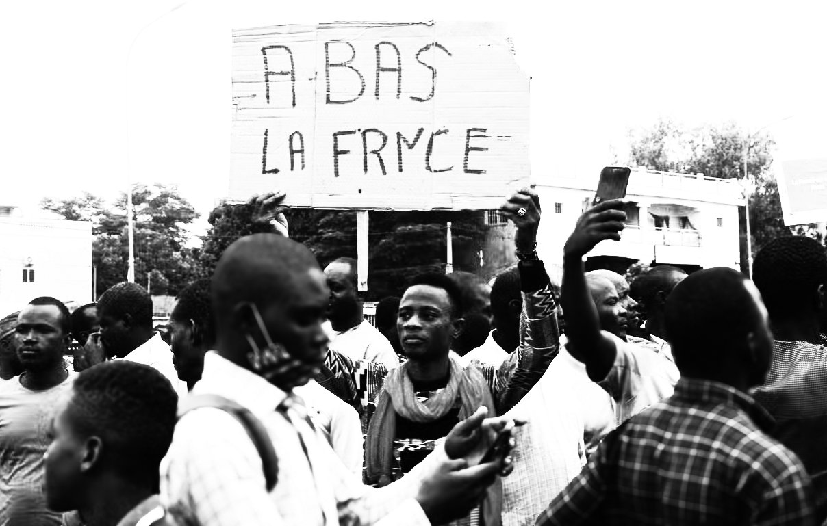 L'image montre une scène de manifestation. Au centre, une personne se tient debout, brandissant une grande pancarte sur laquelle est écrit "À BAS LA FRANCE". Cette phrase semble exprimer un sentiment de mécontentement. Autour de cette personne, un groupe d'hommes et de femmes montre diverses expressions, allant de la détermination à la colère. Certains tiennent des téléphones portables, prenant des photos ou des vidéos de la manifestation. L'arrière-plan est flou, mais on peut distinguer des bâtiments et des arbres. L'atmosphère générale dégage une forte émotion et un engagement collectif sur un sujet qui préoccupe les manifestants.