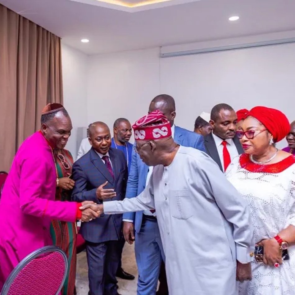 L'image montre un groupe de personnes rassemblées dans une salle, probablement lors d'un événement formel. Au centre, un homme en tenue traditionnelle serre la main d'un autre homme, qui porte une chemise claire. Les autres participants, habillés de manière variée, se tiennent autour d'eux. Certaines femmes portent des vêtements colorés et des accessoires élégants. L'ambiance semble conviviale et respectueuse, avec des sourires et des gestes amicaux. Les murs de la salle sont de couleur claire, et il y a des chaises rouges disposées autour de tables.