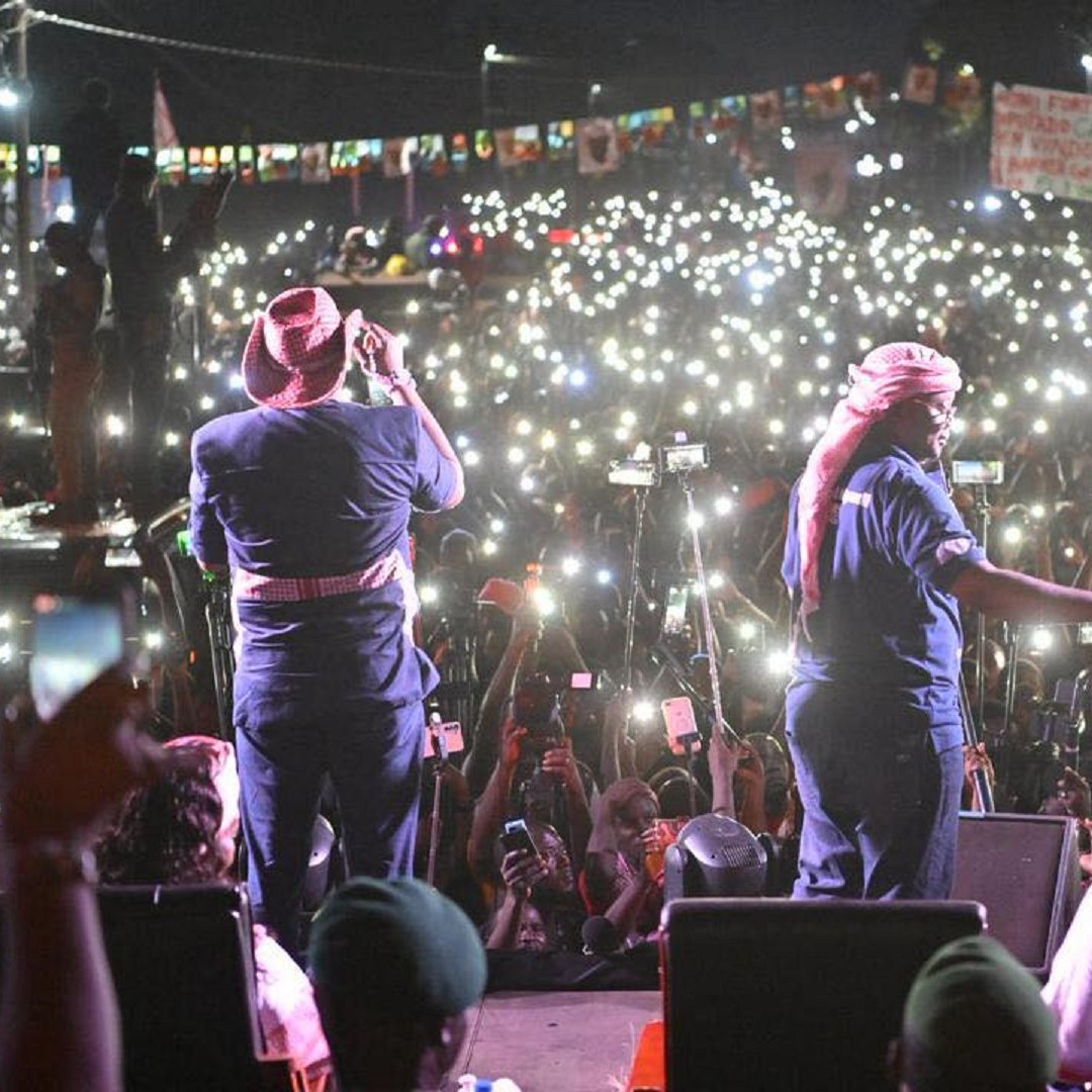 L'image montre une scène d'un concert ou d'un rassemblement en plein air, où un grand nombre de personnes est rassemblé devant un podium. Au premier plan, deux artistes sont sur scène, l'un chantant et l'autre en mouvement, tous deux vêtus de vêtements décontractés, avec des chapeaux et des bandanas. Derrière eux, une mer de spectateurs se tient en arrière-plan, illuminée par d'innombrables lumières de téléphones portables levés, créant un effet scintillant dans la nuit. L'atmosphère est électrisante, avec des couleurs vives provenant des vêtements des membres du public et des affiches. Des banderoles et des drapeaux sont également visibles, ajoutant à l'ambiance festive et engagée de l'événement.