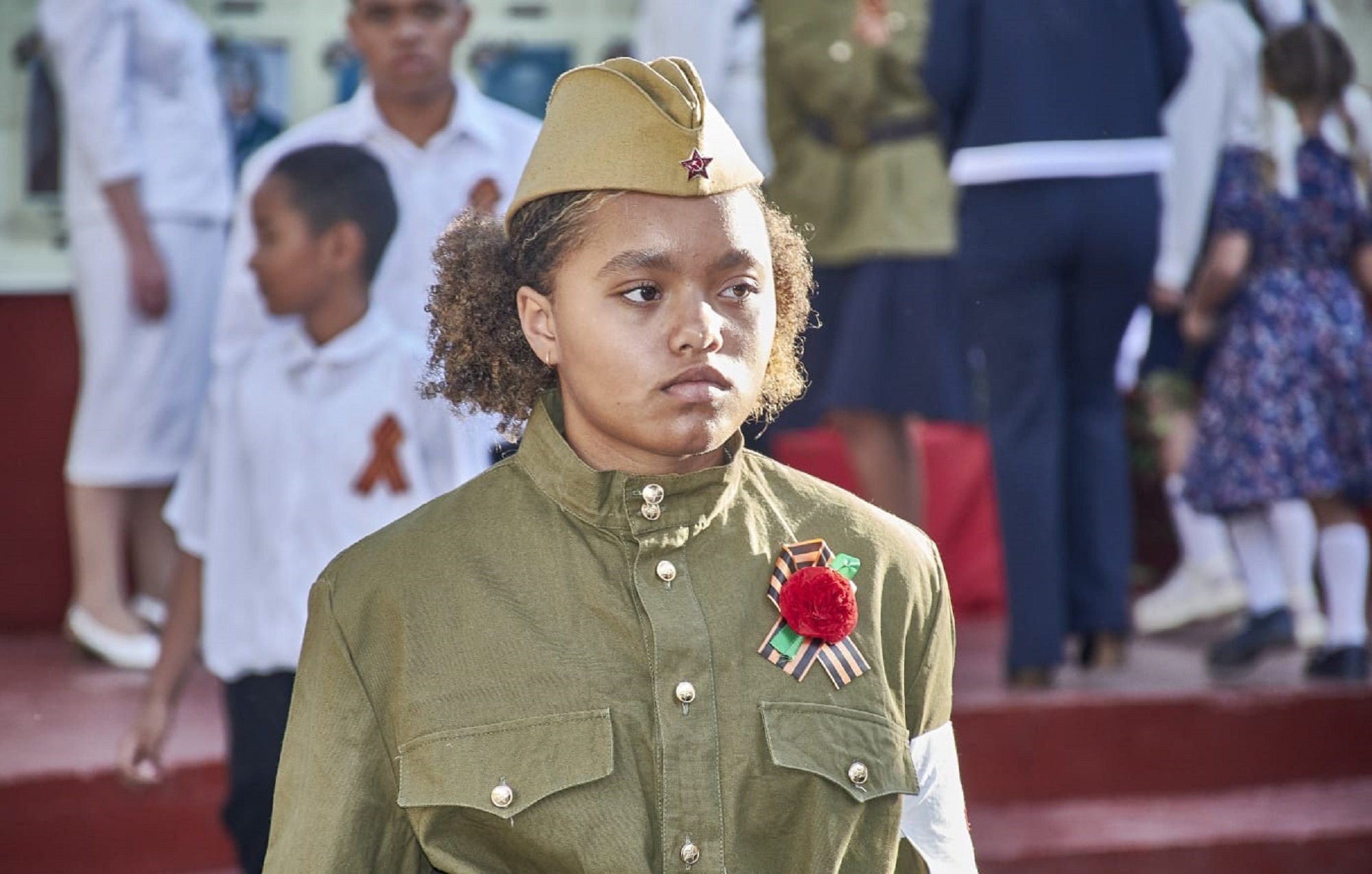 The image features a young girl wearing a military-style uniform with a khaki color. She has a military hat adorned with a star. The uniform includes decorative items like a red flower and a ribbon, possibly indicating a celebration or commemorative event. In the background, there are other children and adults dressed in formal attire, suggesting a public gathering or ceremony, likely related to a historical event. The setting appears to be outdoors, with a backdrop of steps and other participants.