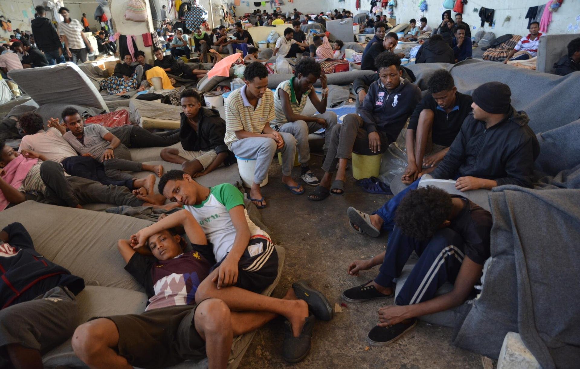 L'image montre un grand espace intérieur, probablement une salle ou un centre d'accueil, où de nombreuses personnes sont rassemblées. Le sol est recouvert de matelas ou de couvertures, et les gens sont assis ou allongés, souvent en petits groupes ou seuls. Les visages montrent une variété d'émotions, allant de la fatigue à l'attente. L'atmosphère semble chargée, et l'agencement des personnes donne une idée d'une situation difficile. Les murs sont nus, et il y a des effets personnels éparpillés ici et là. L'environnement est sombre et témoigne d'un manque de confort.