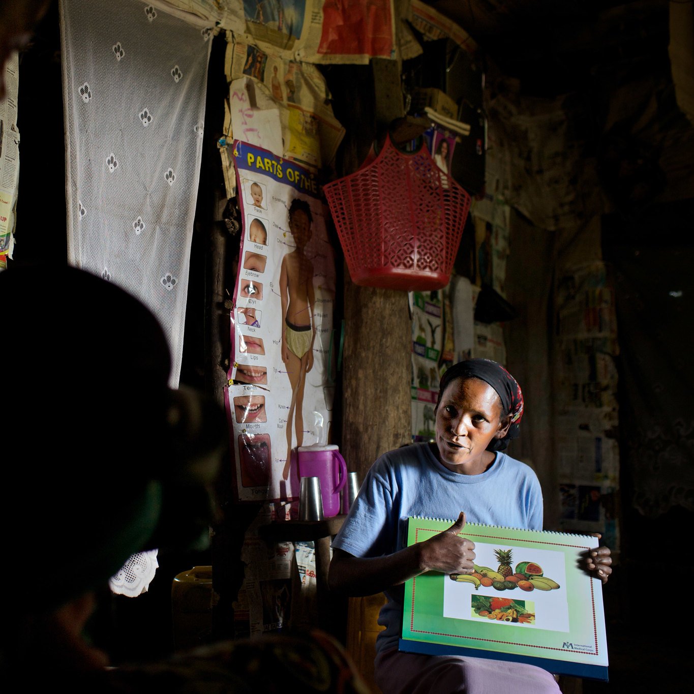 L'image montre une femme assise dans une pièce sombre, entourée de murs décorés d'affiches et de tissus. Elle tient un grand carnet avec des illustrations de fruits, qu'elle montre avec enthousiasme. Sa tenue est simple, et elle a l'air concentrée sur son explication. À l'arrière-plan, une silhouette d'une autre personne est visible, ajoutant à l'atmosphère d'échange. La scène évoque un moment d'apprentissage ou de partage d'informations sur les fruits.
