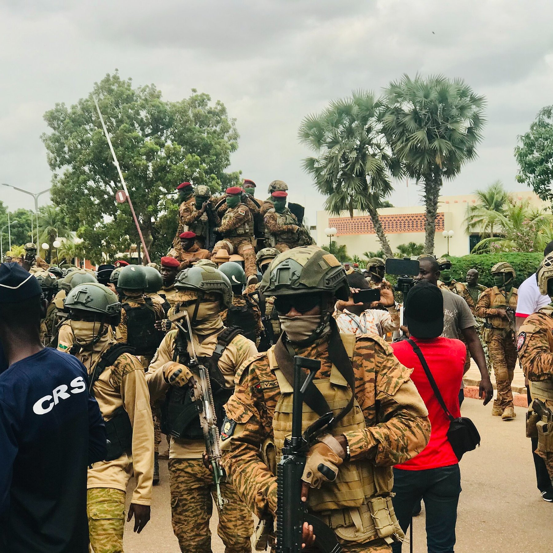The image depicts a scene of armed soldiers in a public area. The soldiers are wearing camouflage uniforms and helmets, some with face coverings, and are equipped with rifles. In the background, there are additional soldiers on a vehicle. The atmosphere appears to be tense, with a gathering of civilians present as well, some taking photographs. There are trees and buildings in the background, suggesting an urban setting. Overall, the scene conveys a strong military presence.