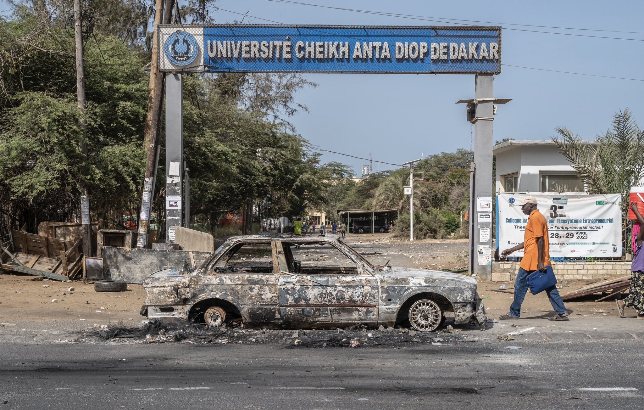 L'image montre l'entrée de l'Université Cheikh Anta Diop de Dakar, identifiable par le panneau au-dessus. En premier plan, une voiture calcinée est garée sur la route, avec des traces de brûlures et des dégâts visibles, indiquant qu'elle a été abandonnée après un incendie. En arrière-plan, on peut apercevoir des arbres et des structures, mais l'atmosphère semble marquée par un sentiment de désolation. Un piéton, portant une sacoche, marche sur le trottoir, ajoutant un contraste entre l'activité humaine et l'état dégradé des lieux.
