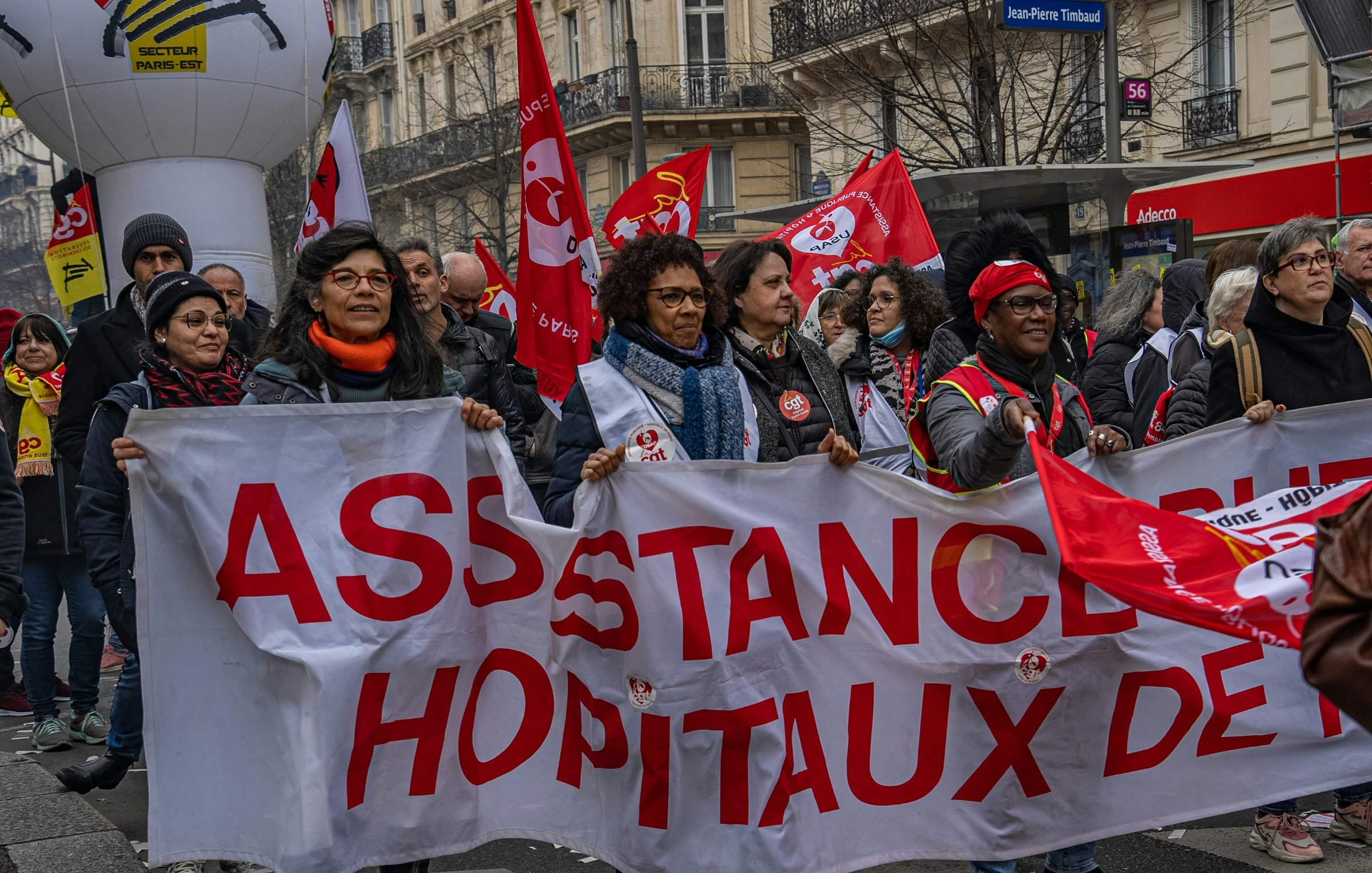 L'image montre une manifestation dans une rue animée. Au premier plan, un groupe de personnes tient une grande banderole blanche sur laquelle est écrit "ASSISTANCE HÔPITAUX DE…" en lettres rouges. Les manifestants arborent des sourires et semblent déterminés. On aperçoit également plusieurs drapeaux rouges, qui sont souvent associés à des syndicats. L'atmosphère est dynamique, avec une multitude de personnes autour, représentant la solidarité et l'engagement. Les bâtiments en arrière-plan suggèrent une ambiance urbaine, typique des grandes villes.