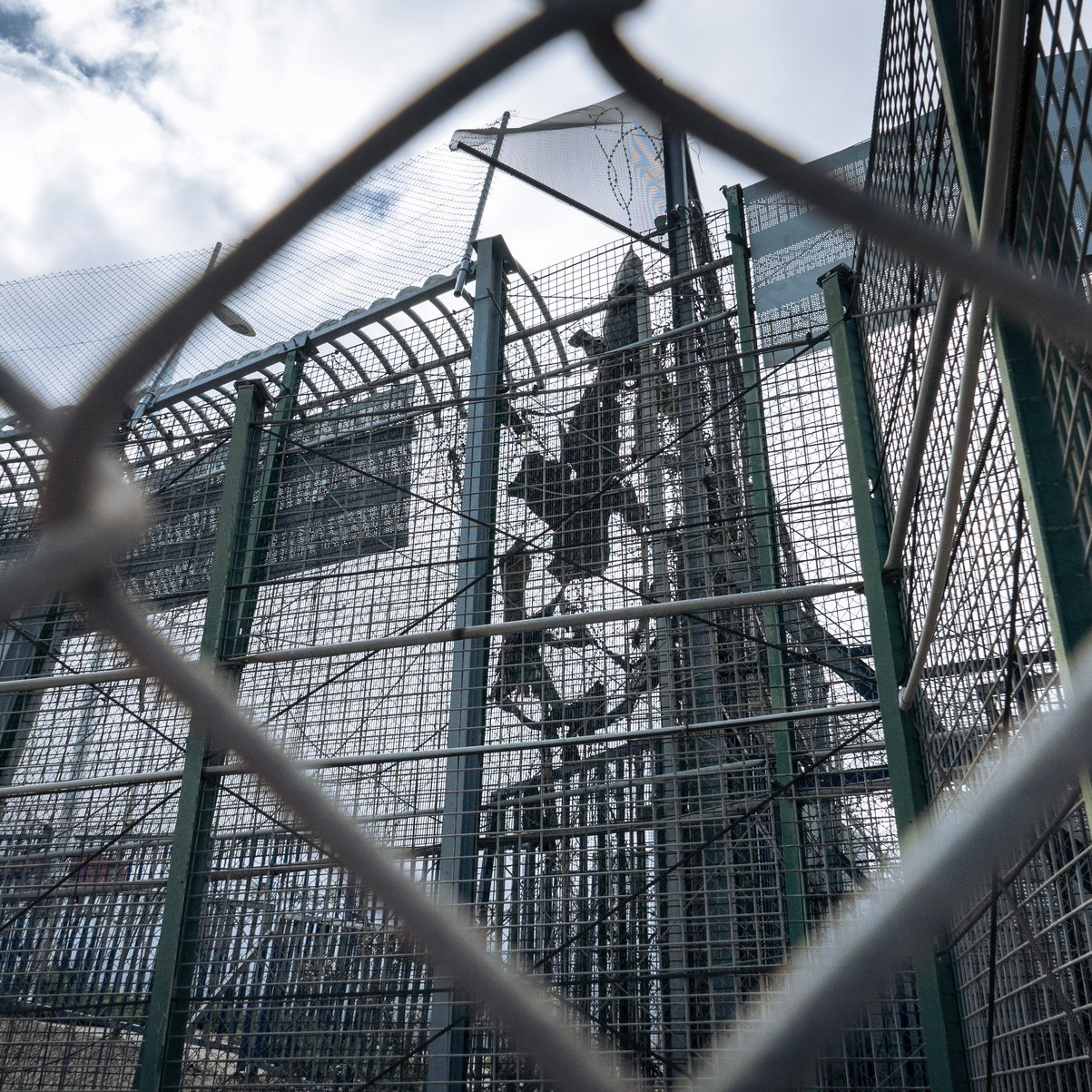 L'image représente une structure de sécurité, probablement une clôture ou un mur de protection. On aperçoit des barrières métalliques en grillage, qui sont transparentes et permettent de voir à travers. À l'intérieur, on distingue une installation, probablement en hauteur, marquée par des éléments solides tels que des poteaux. Le ciel en arrière-plan est nuageux, laissant entendre une lumière diffuse. L'ensemble dégage une impression d'isolement et de confinement, accentuée par la perspective de la clôture.