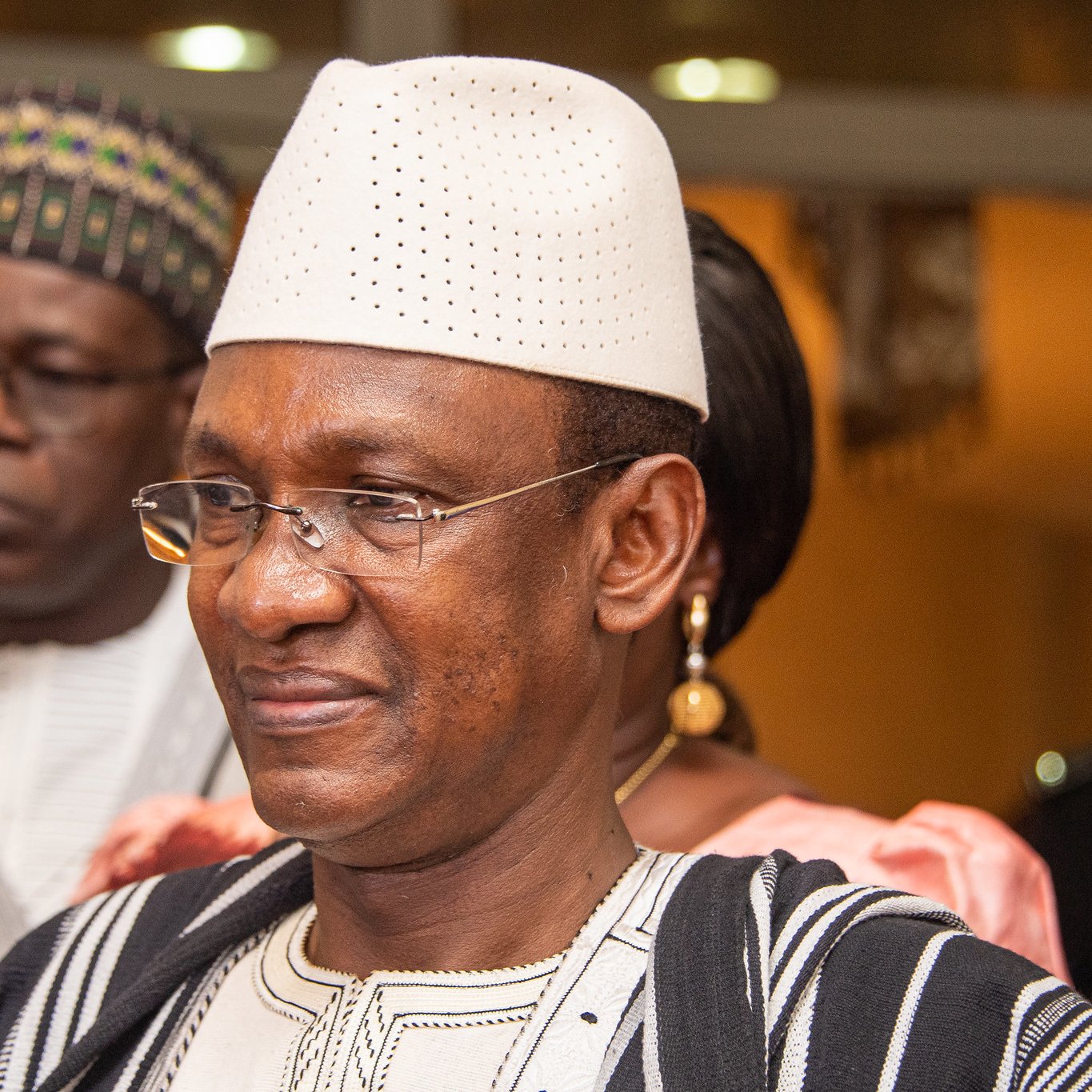 Dans cette image, nous voyons un groupe de personnes dans un cadre formel. L'homme au premier plan porte un chapeau traditionnel blanc et une tenue en tissu bicolore, avec des motifs caractéristiques. Il a un sourire serein sur le visage, illustrant une certaine dignité et bienveillance. À l'arrière-plan, deux autres personnes sont présentes, l'une portant des lunettes et l'autre en tenue traditionnelle également. L'éclairage de la scène suggère une ambiance chaleureuse et accueillante, typique d'un événement social ou culturel.