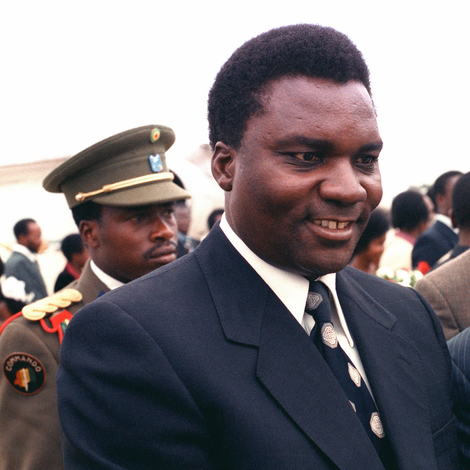 L'image montre un groupe de personnes en extérieur lors d'un événement formel. Au premier plan, deux hommes bien habillés sourient et semblent discuter. L'un porte un costume noir avec une cravate à motifs, tandis que l'autre arbore un costume rayé avec une cravate aux motifs plus délicats. En arrière-plan, un homme en uniforme militaire observe la scène, ce qui ajoute une dimension de solennité à l'événement. Le fond est lumineux, et on peut deviner une atmosphère de convivialité et d'échange entre les participants.
