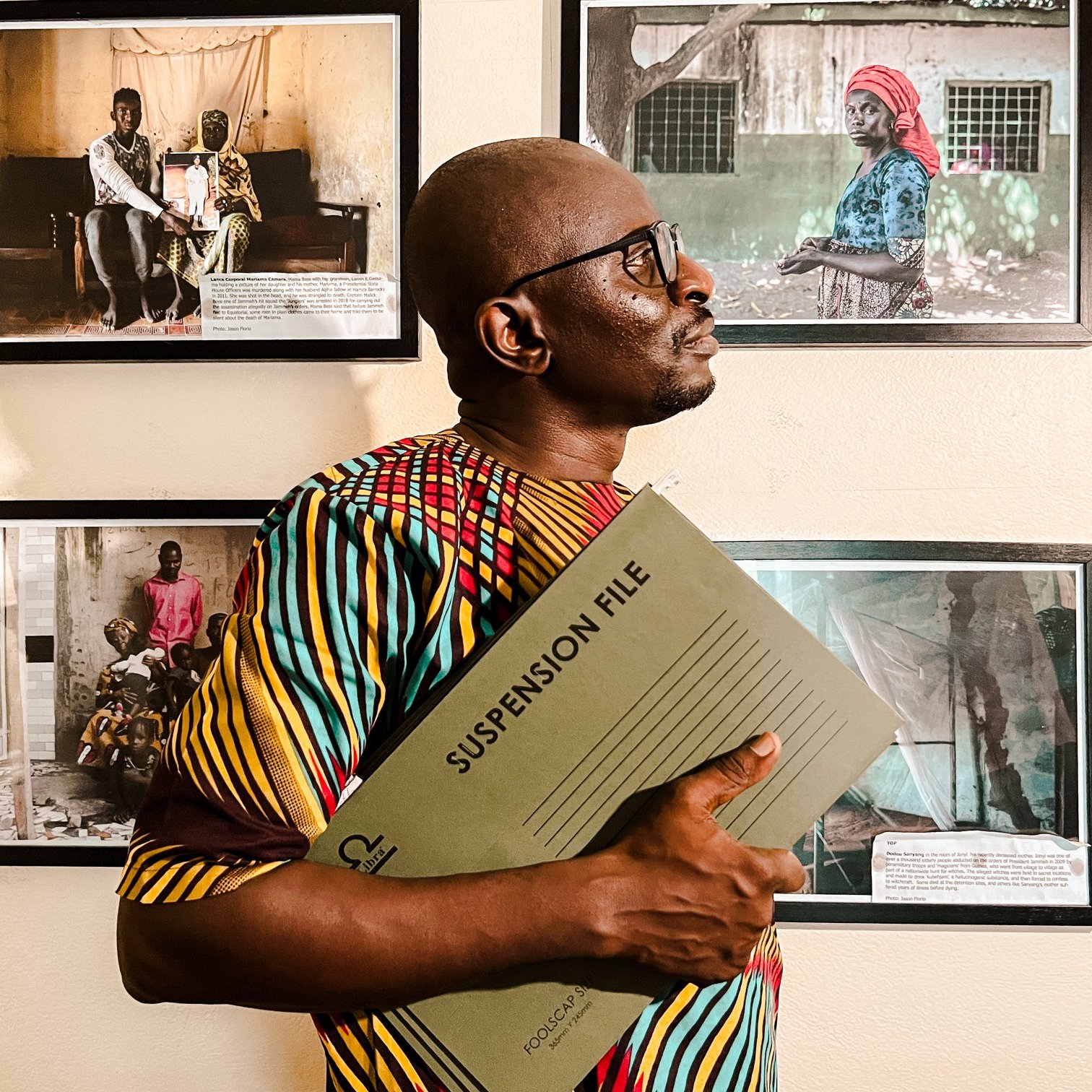 L'image montre un homme debout dans un espace d'exposition, entouré de plusieurs photographies accrochées au mur. Il porte une chemise colorée aux motifs vibrants, mêlant des teintes de jaune, rouge et vert. Dans ses bras, il tient un dossier intitulé "SUSPENSION FILE", ce qui suggère qu'il se prépare à discuter ou à présenter quelque chose d'important. L'homme se tourne légèrement sur le côté, regardant vers les photos, qui semblent raconter des histoires de vie, avec des portraits d'autres personnes. L'atmosphère de l'image est à la fois sérieuse et engagée, soulignant l'importance des histoires capturées dans ces œuvres visuelles.