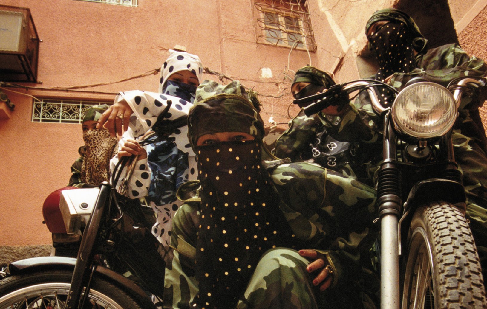 L'image montre un groupe de personnes habillées de manière distinctive, portant des vêtements camouflés, ainsi que des masques et des foulards ornés de paillettes ou de motifs. Ils se trouvent dans une rue d'une vieille ville, probablement dans un environnement urbain. À côté d'eux, deux motos ajoutent un élément de modernité à la scène. Leurs poses évoquent une certaine solidarité et un esprit de camaraderie. L'éclairage est naturel, suggérant un moment de la journée typique dans une ville animée. Les textures des vêtements contrastent avec le pavé de la rue, créant une ambiance unique.