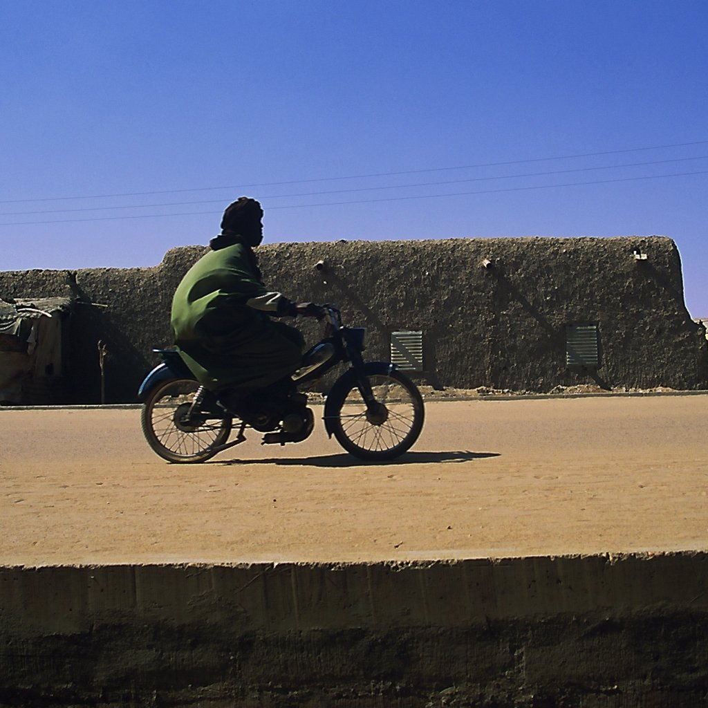 L'image montre un homme en train de rouler à bicyclette sur une route en terre. Il porte une tenue traditionnelle de couleur verte, qui contraste avec le décor. En arrière-plan, on peut apercevoir des bâtiments en adobe, typiques des régions désertiques, avec des murs de terre et des fenêtres. Le ciel est d'un bleu éclatant, sans nuages, et l'atmosphère générale est calme, suggérant une journée ensoleillée. La lumière du soleil éclaire la scène, mettant en valeur les textures du sol et des constructions.