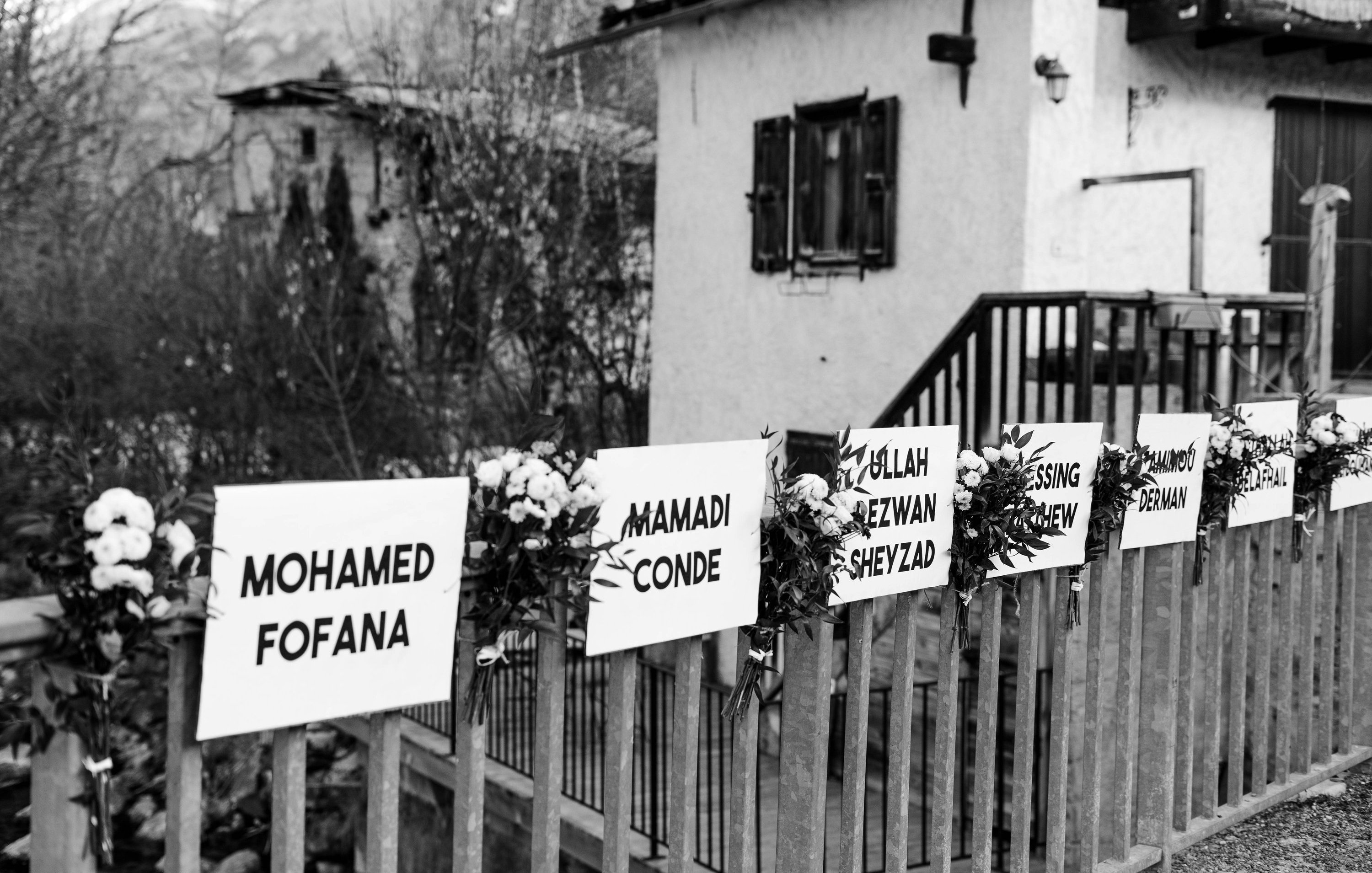 L'image montre une scène en noir et blanc, où l'on aperçoit une maison avec un balcon en bois. Devant la maison, il y a une clôture sur laquelle sont fixés plusieurs panneaux blancs. Chaque panneau présente un prénom et un nom, probablement en hommage à des personnes. Des fleurs blanches sont disposées autour de chaque panneau, ajoutant une touche de douceur. En arrière-plan, on peut deviner des arbres et la nature environnante, suggérant un cadre paisible et naturel.